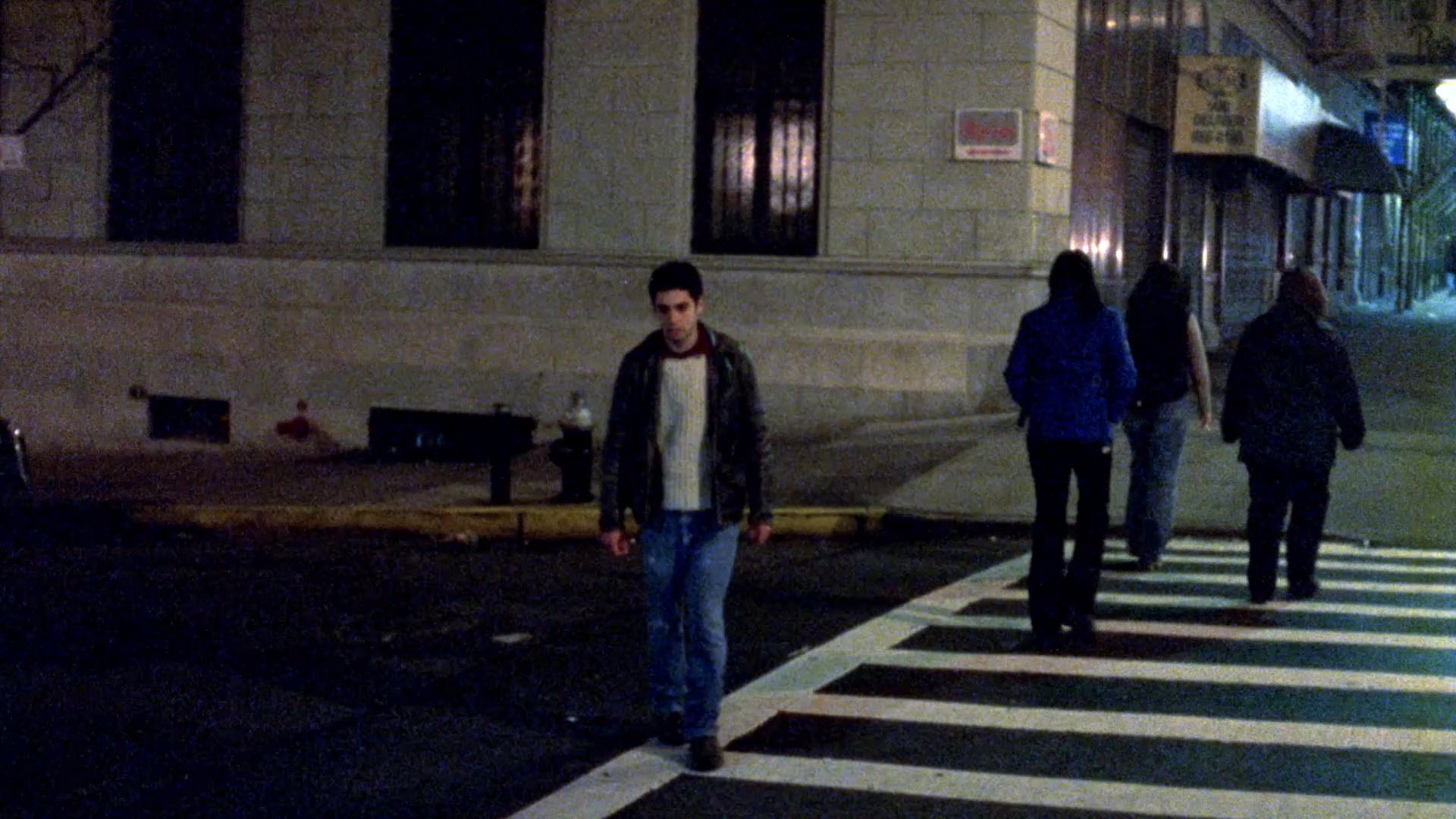 A man in a leather jacket traverses a city street at night, following a group of people before moving into a park area. The sequence blends urban realism with abstract animated effects during a period of movement.