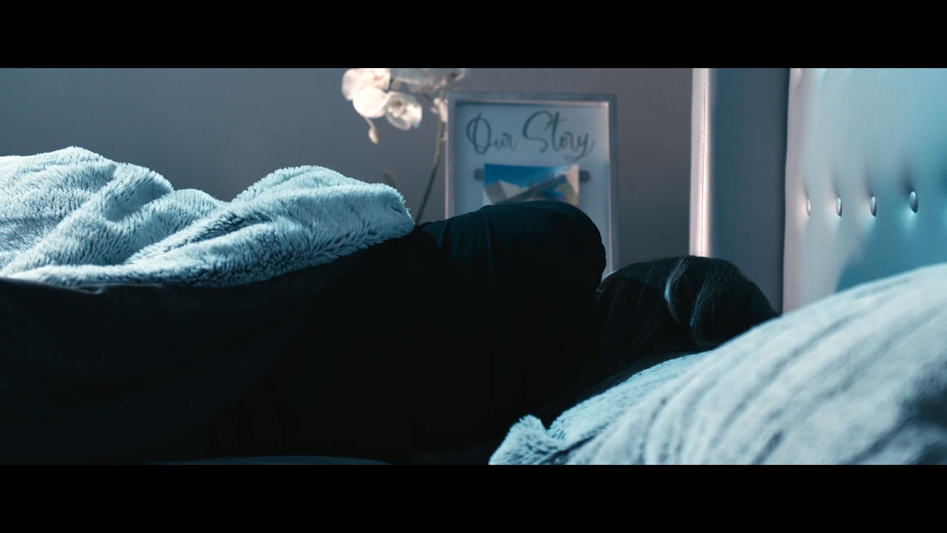 A woman lies in bed, stirring and waking from sleep in a dimly lit bedroom environment.