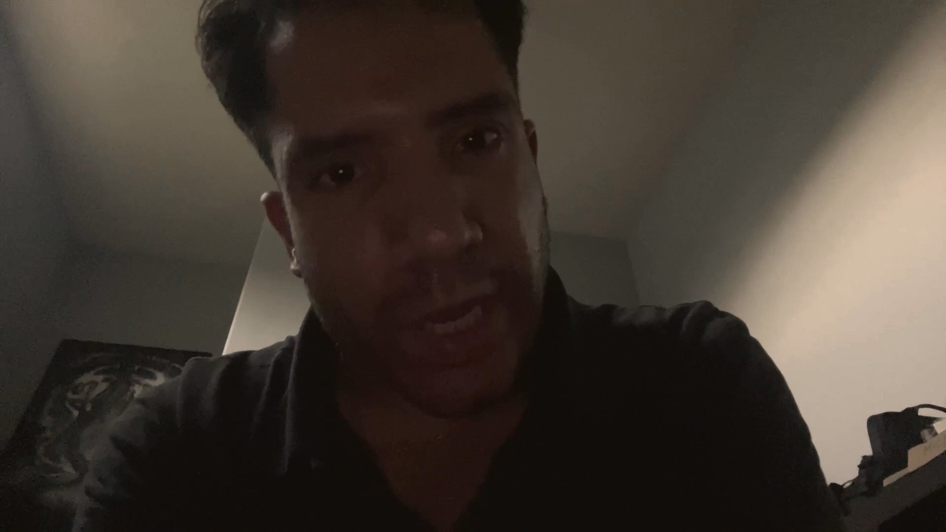 A man addresses the camera directly in a dimly lit, casual indoor setting, delivering a personal monologue.