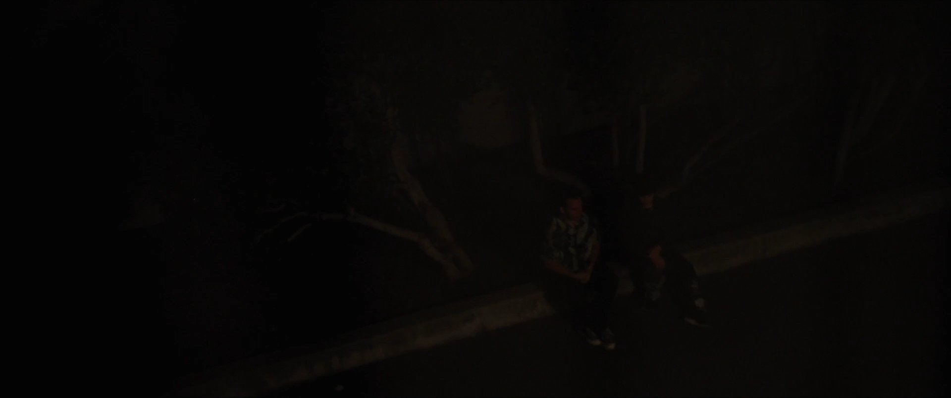 Two individuals engage in a tense conversation at night, while a third person watches silently from a lit balcony above.
