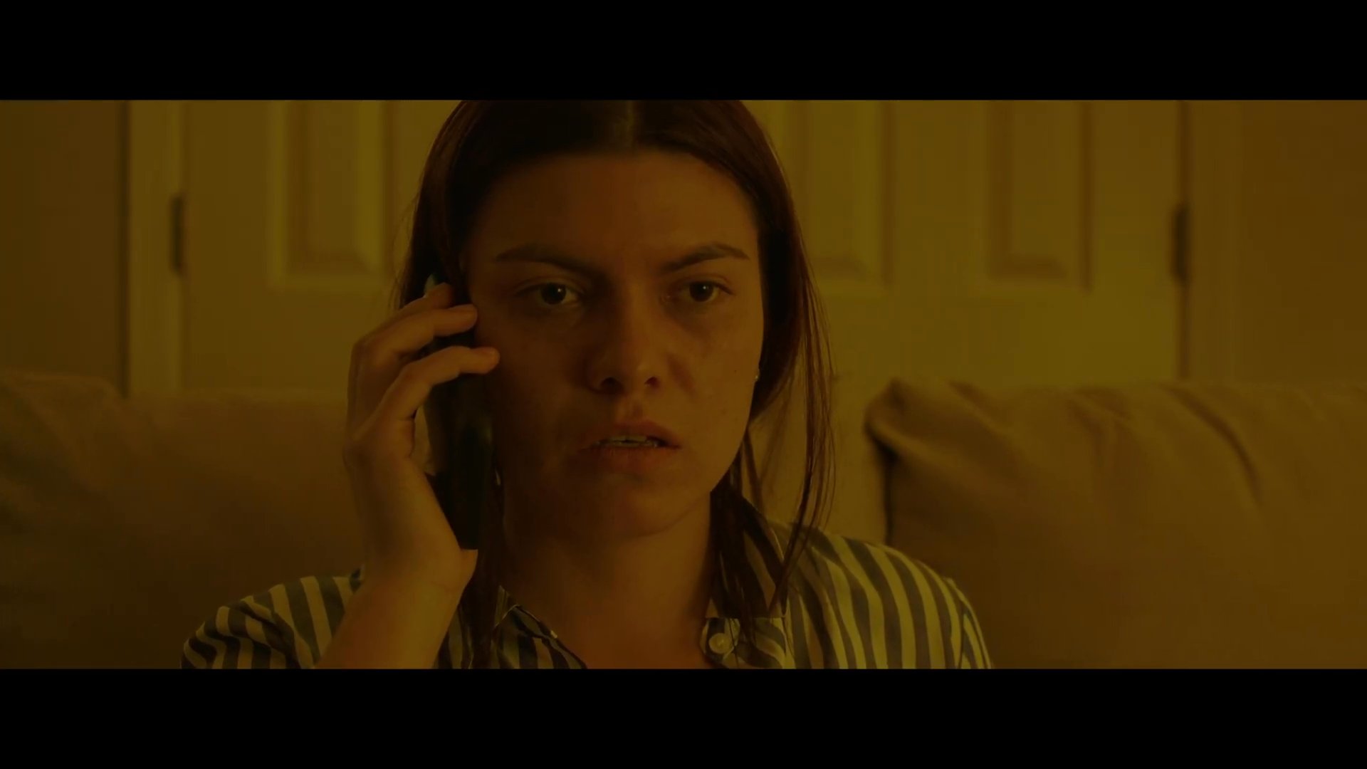 A woman sits on a sofa with her daughter and experiences visible emotional distress while engaged in a somber telephone call. The scene conveys a sense of mounting tension and concern within a domestic environment.