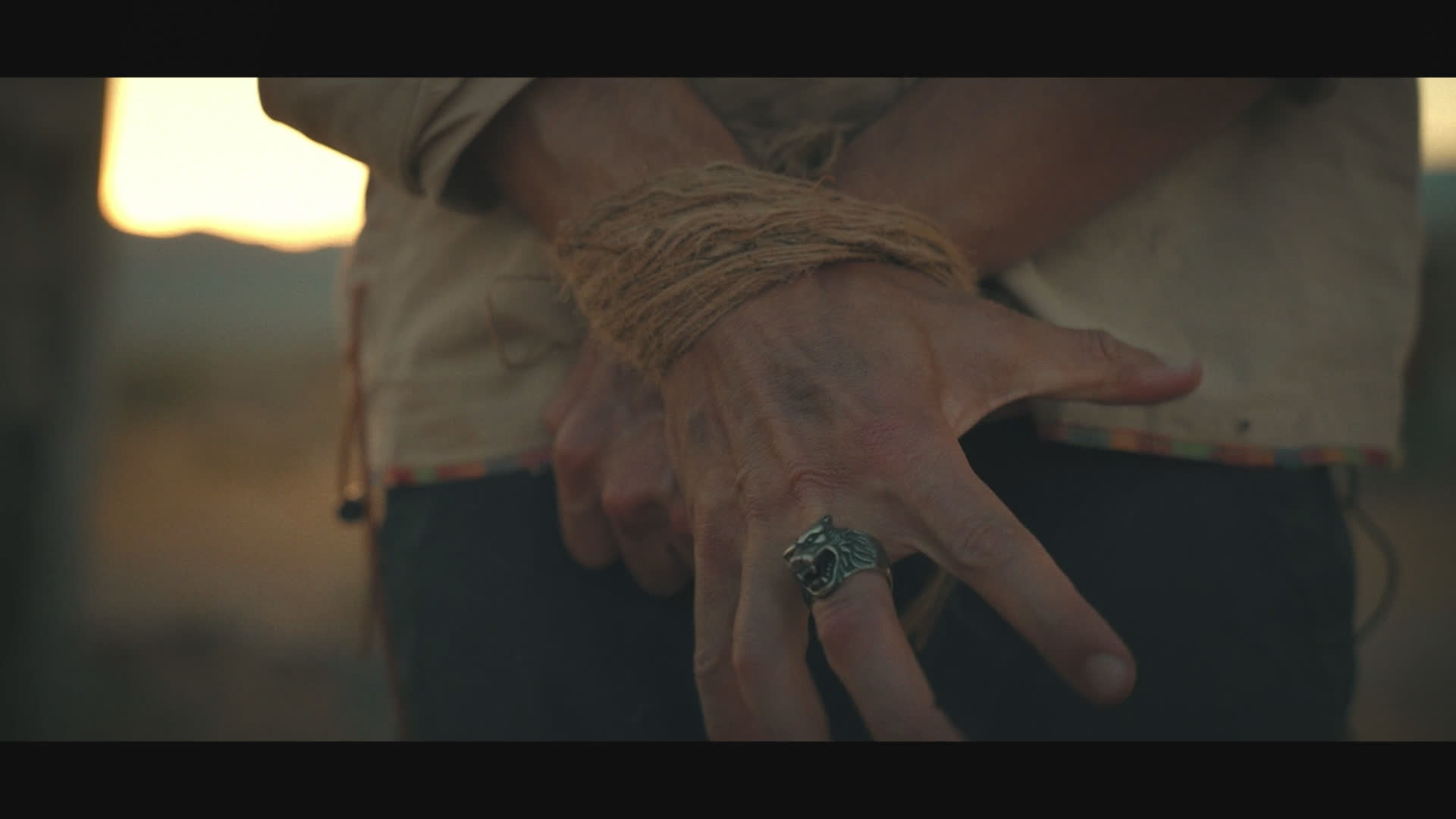 A solitary settlement sits beneath a water tower as a figure presses a limb into a debris-filled basin. Close-ups reveal bound wrists and a long-haired man whose tense gaze shifts across the barren landscape.