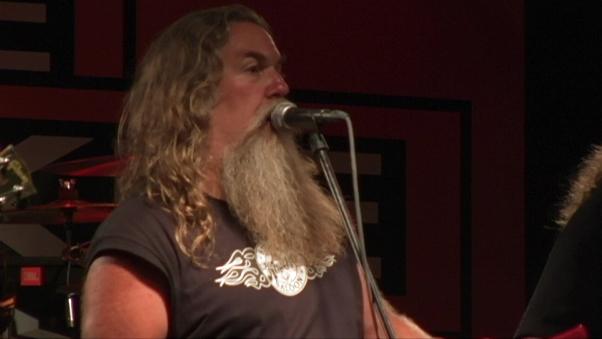 A long-bearded musician transitions from performing on stage with a guitar to riding a motorcycle through a forest at sunset. This visual contrast highlights a lifestyle blend of musical artistry and adventurous travel.