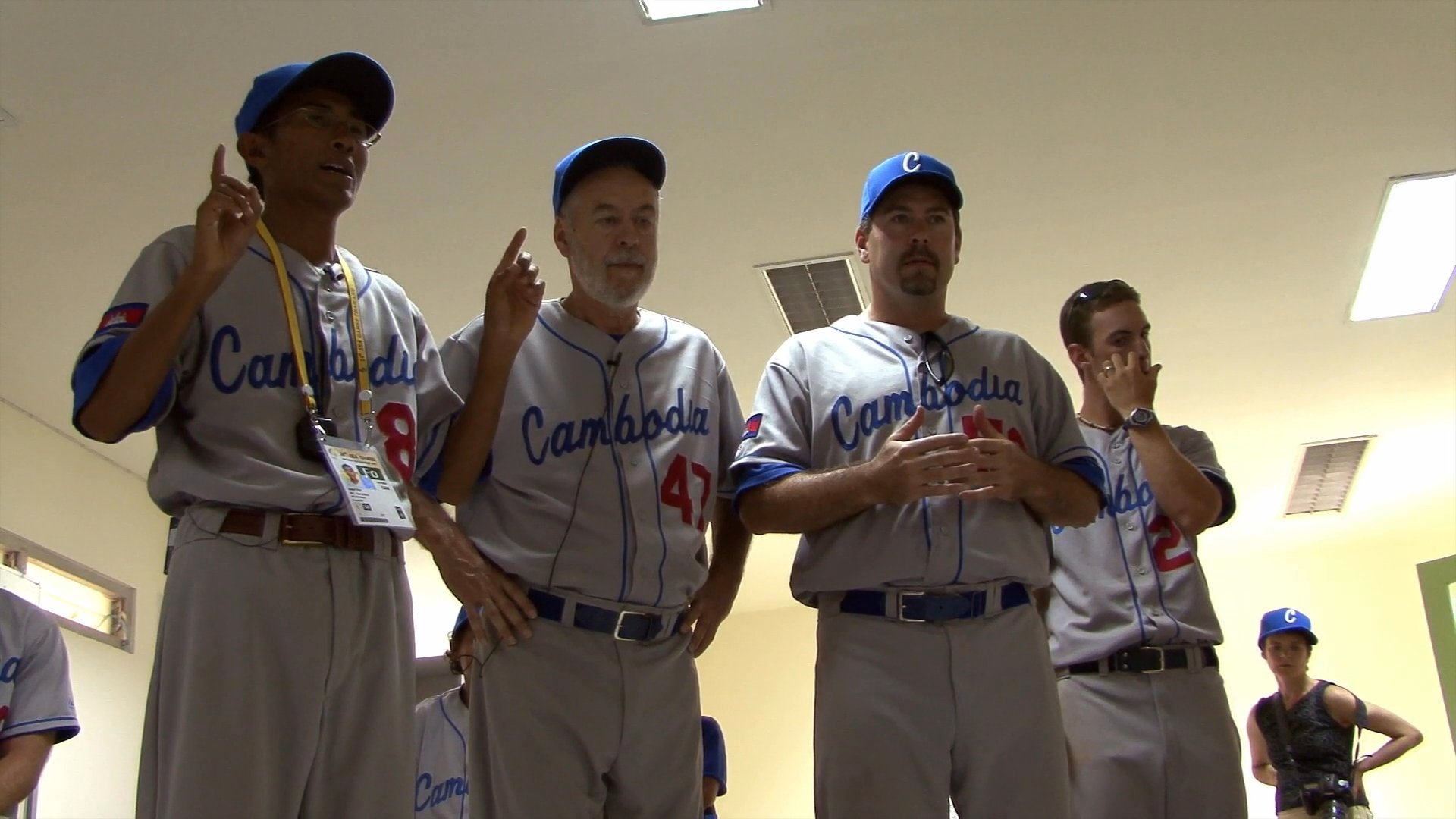Members of the Cambodia baseball team gather in a locker room setting, listening intently as they prepare for an upcoming event or game. The group exhibits a spirit of professional camaraderie and focus while dressed in their team uniforms.