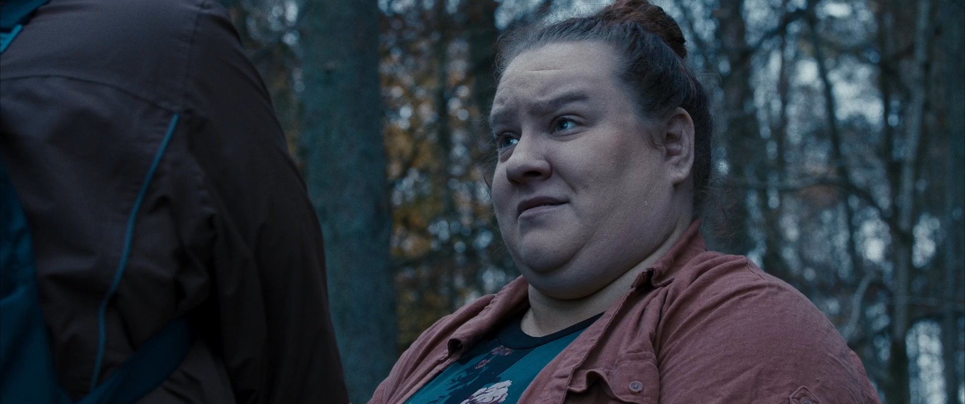 Two individuals engage in a conversation while walking through a dim forest, eventually arriving at a secluded cabin.