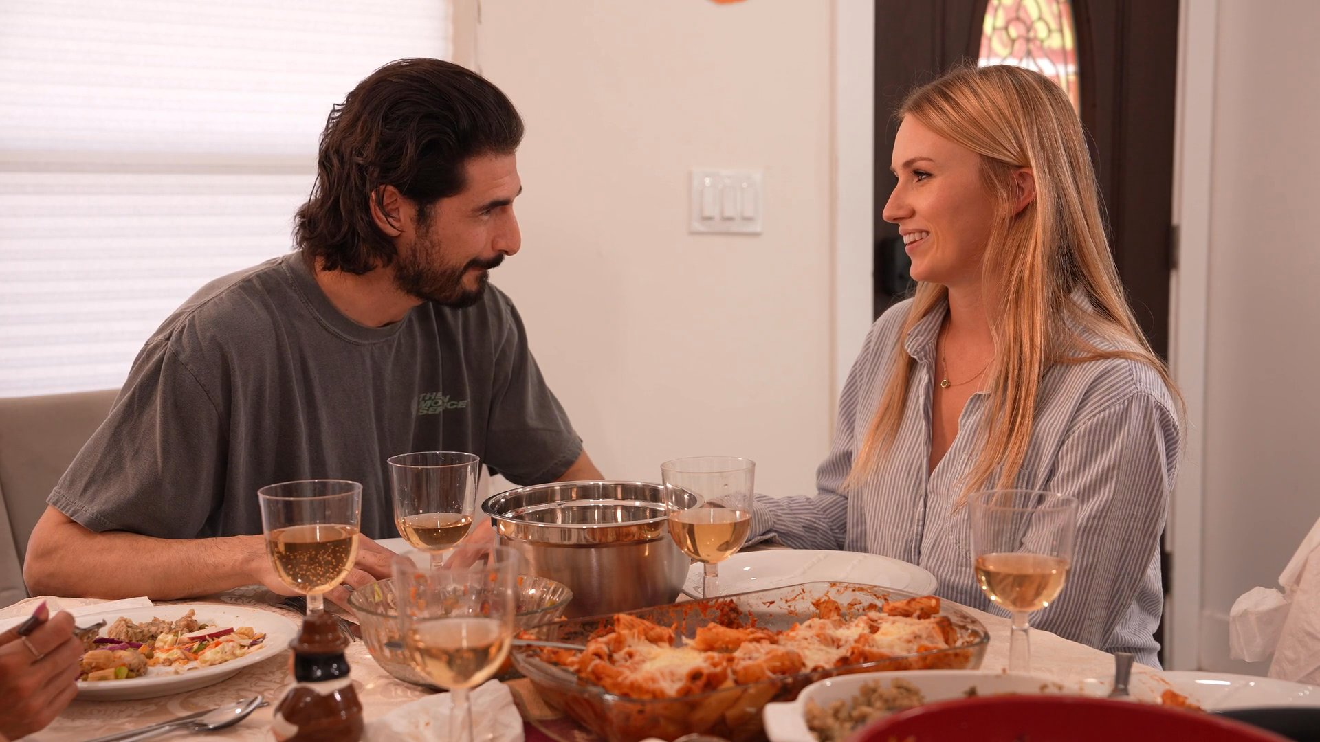 A man and a woman engage in conversation while seated at a dining table filled with food and drinks. The setting suggests an intimate domestic meal shared between the two individuals.