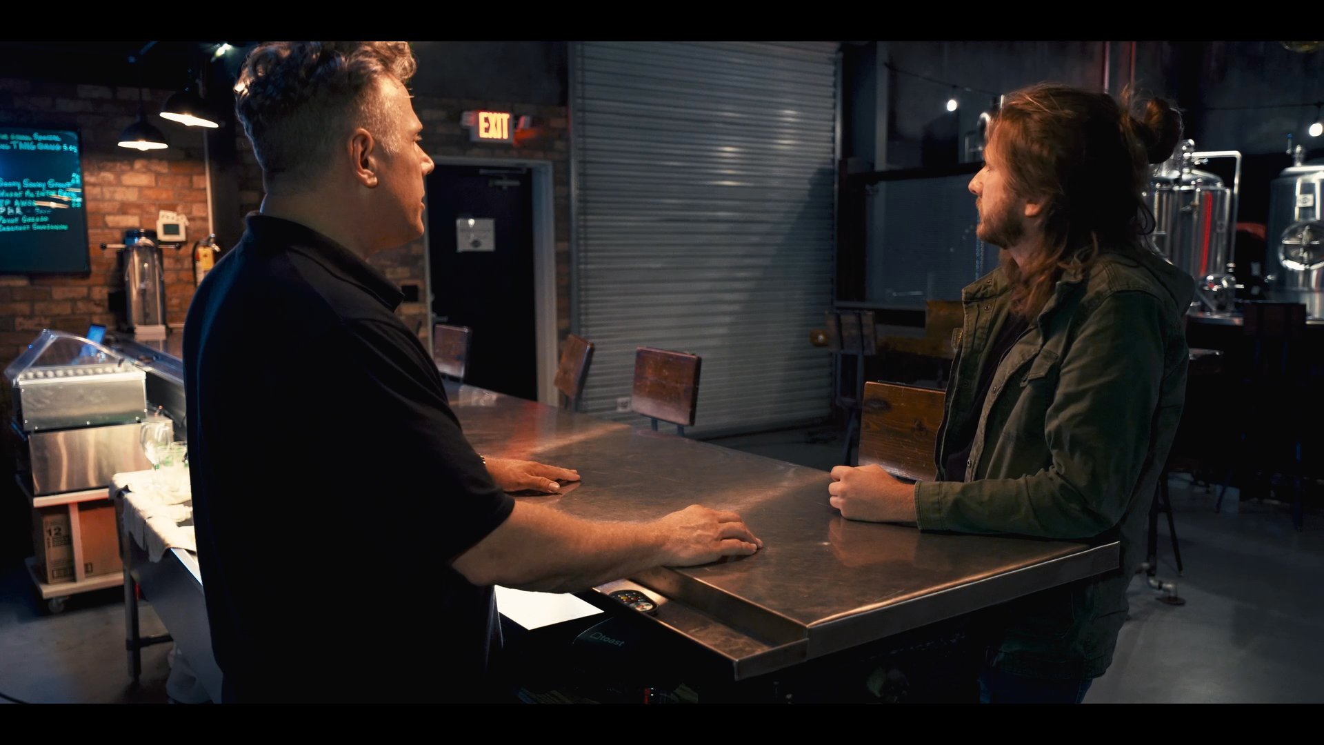 Two individuals engage in a professional discussion while standing at a stainless steel table within a brewery environment. The exchange focuses on collaborative communication and project planning.