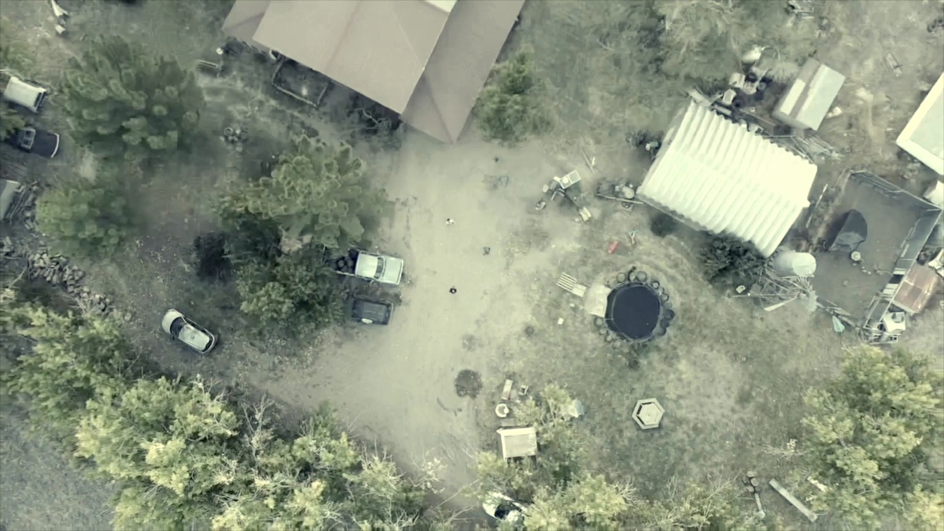 An aerial perspective captures a rural property featuring a residential building, parked vehicles, and surrounding vegetation. The scene provides a topographical view of the homestead setting.