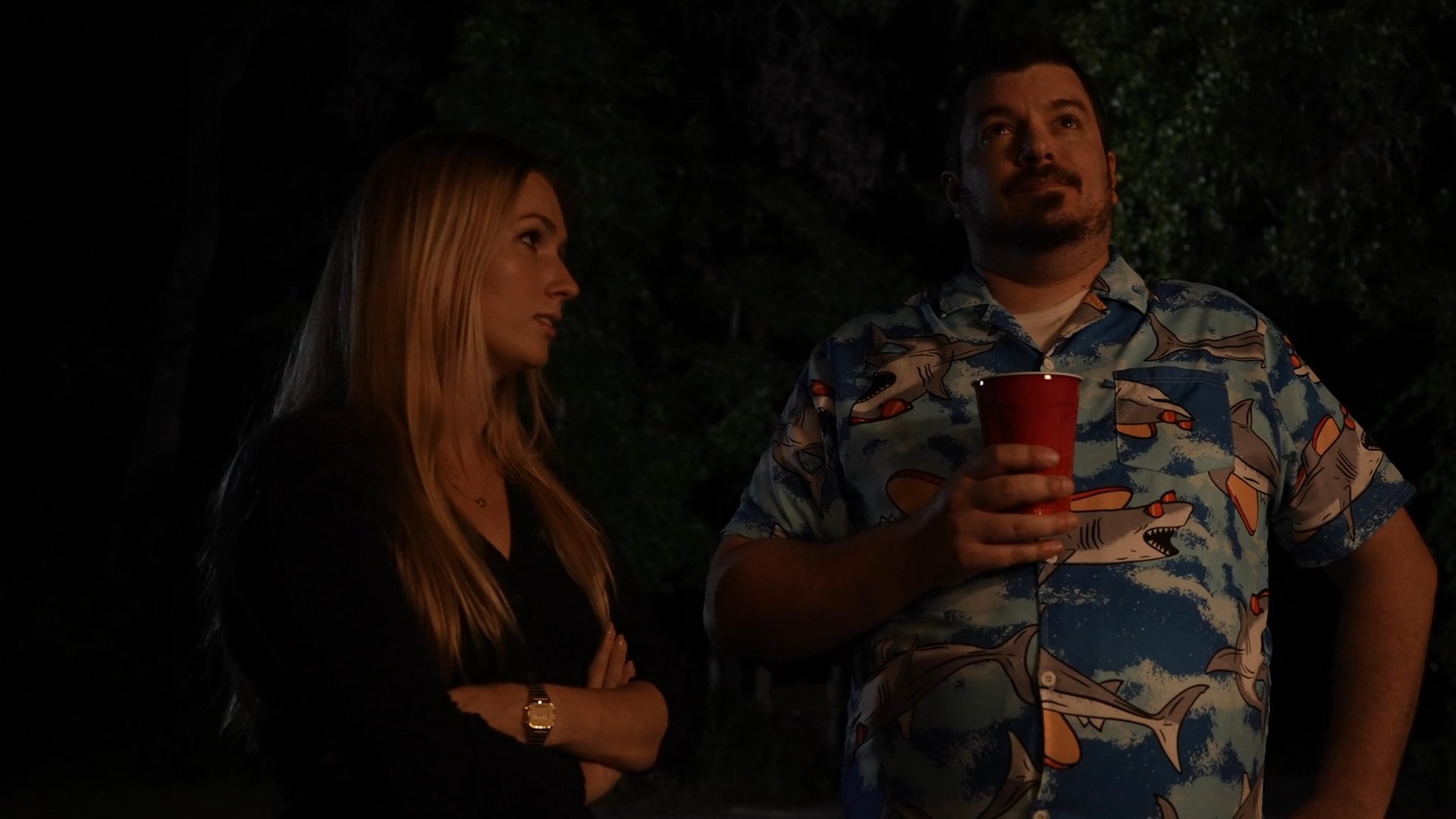 A woman and a man in a patterned shirt stand together during an evening event, looking upward while engaging in a quiet conversation.