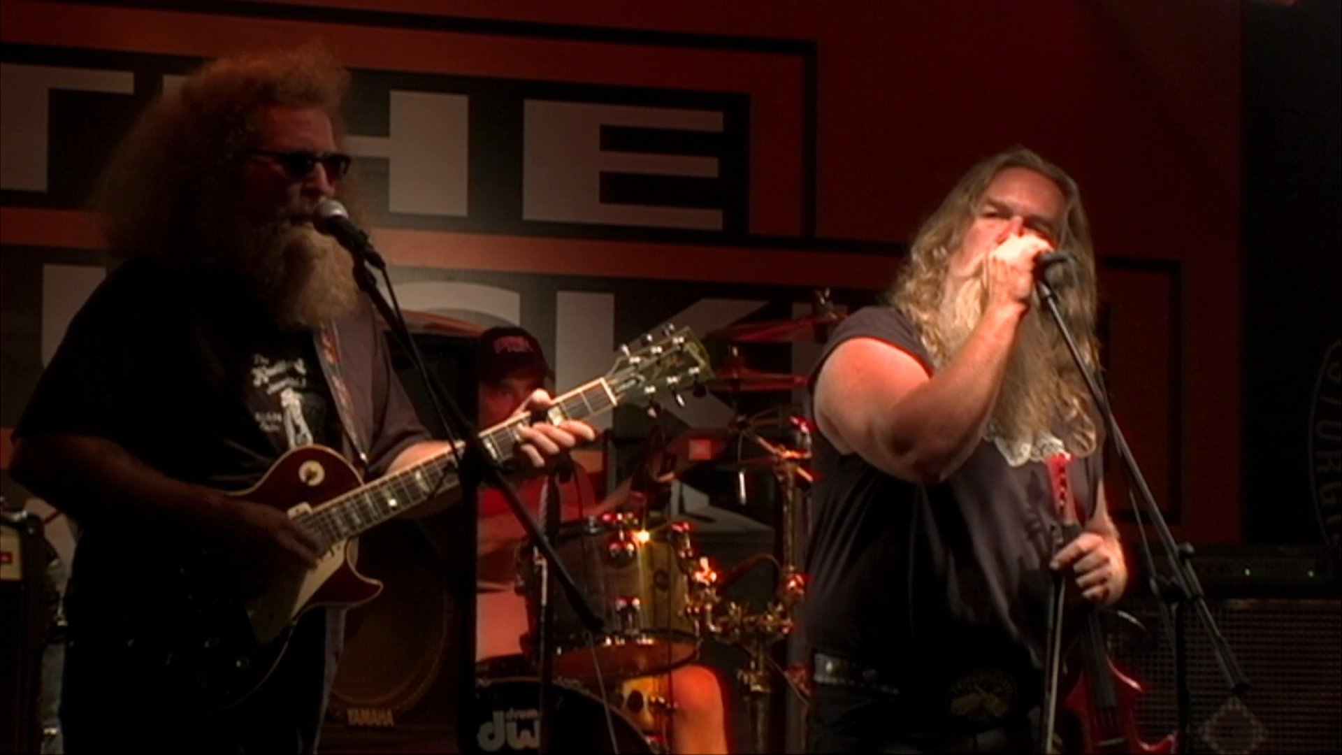 A long-haired, bearded rock musician performs on stage before a lively audience at a dimly lit bar. The atmosphere captures a vibrant nightlife scene with patrons dancing and enjoying the musical performance.