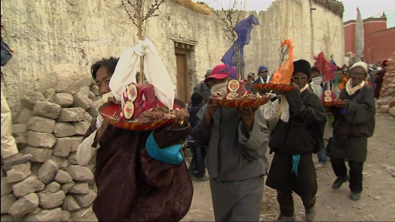 Devotees spin prayer wheels at a Tibetan monastery before joining a colorful religious procession through the village. Participants in traditional attire carry ritual offerings during this communal cultural event.