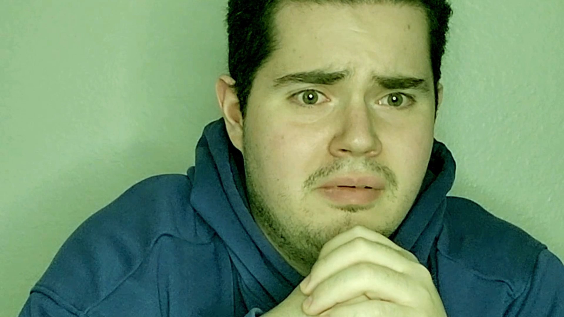 A man exhibits signs of deep contemplation and concern, his facial expressions shifting while he keeps his hands clasped beneath his chin. The minimalist setting keeps the focus entirely on his emotive responses.