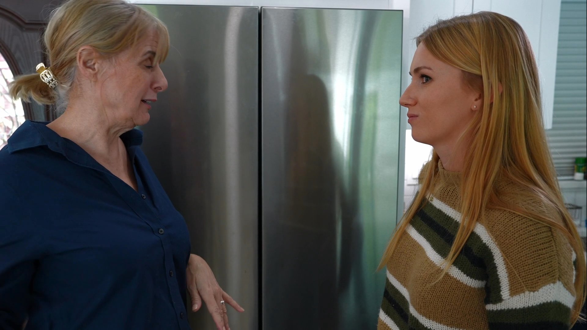 Two women share an emotional reunion in a home kitchen, exchanging a warm embrace followed by an engaging, lively conversation.