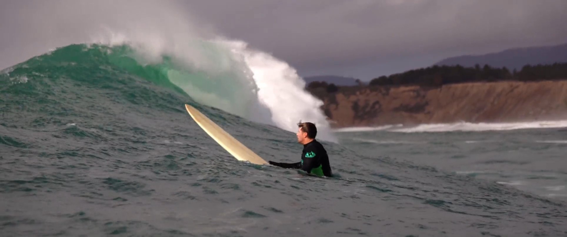 A surfer in a wetsuit paddles through open water while observing incoming waves along a coastal shoreline.