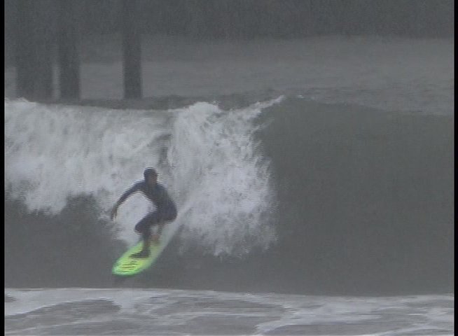 A surfer navigates challenging waves near a coastal pier during inclement weather. The footage captures the athlete executing turns and maneuvering through the surf.