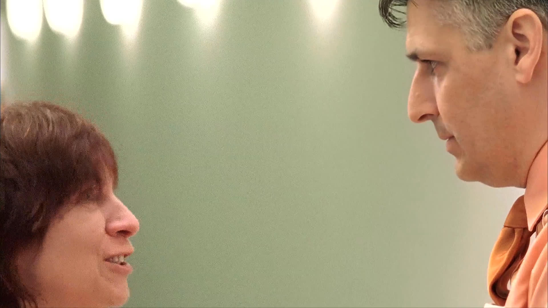 A woman and a man engage in a tense, animated conversation in a brightly lit hallway before the scene transitions to a dimly lit bedroom setting.