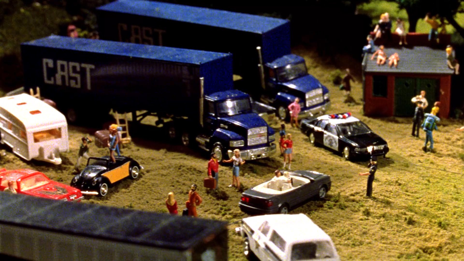 A live musical performance transitions to a detailed diorama depicting a busy film production site with miniature vehicles, crew members, and equipment. The scene highlights the intricate construction of a scale model environment used to represent an outdoor film location.