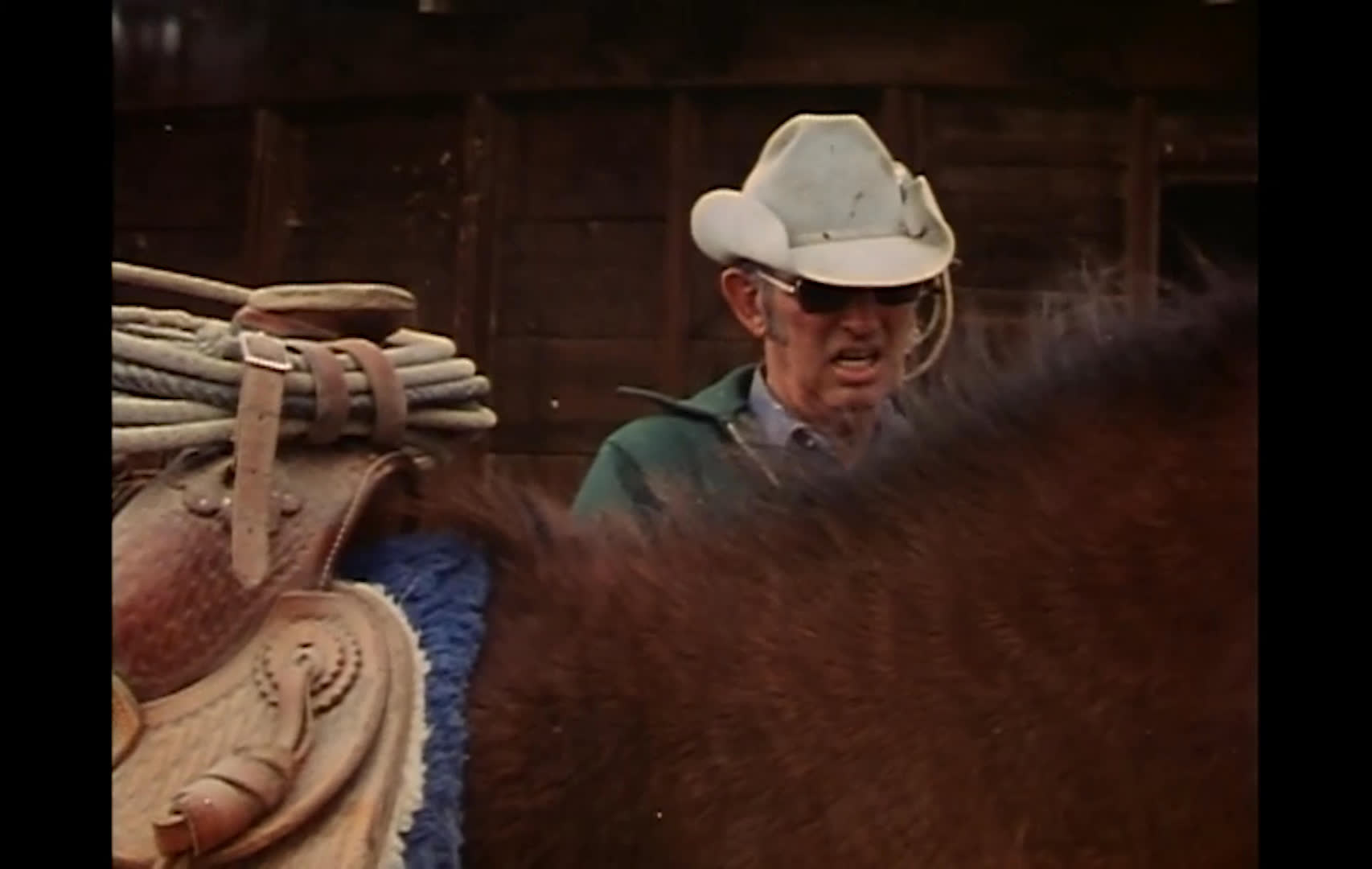 A rancher in a white hat speaks beside a saddled horse at a wooden barn. Cattle graze near a reflective pond beneath a cloudy mountain skyline as he describes the valley’s livestock and hay country.