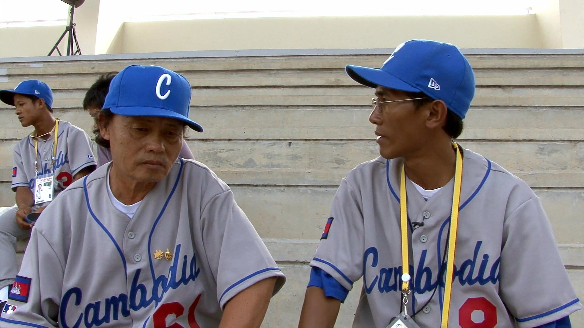Members of the Cambodia national baseball team sit in a stadium setting while participating in an interview regarding their sport and athletic training.
