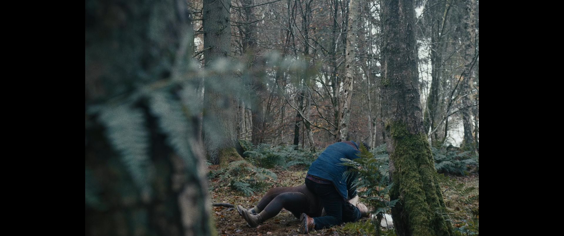 A tense struggle unfolds in a wooded area as an individual is forcibly restrained on the ground by an assailant. The scene conveys high levels of distress and physical conflict.