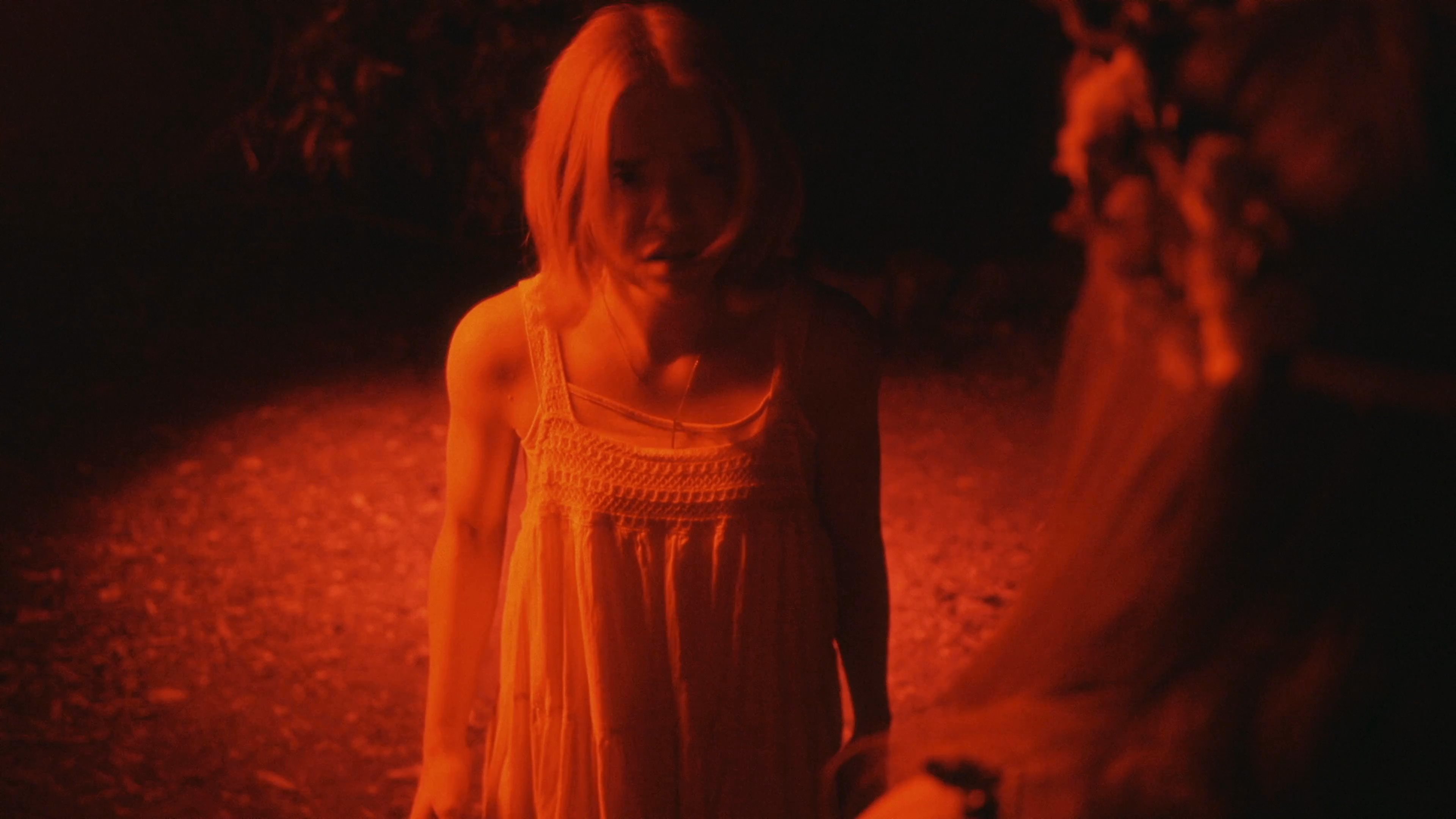 Two women interact in a dimly lit, red-hued forest setting while engaged in a mysterious, ritualistic exchange involving a floral offering. The atmospheric scene emphasizes tension and cryptic storytelling through minimal dialogue and stylized lighting.
