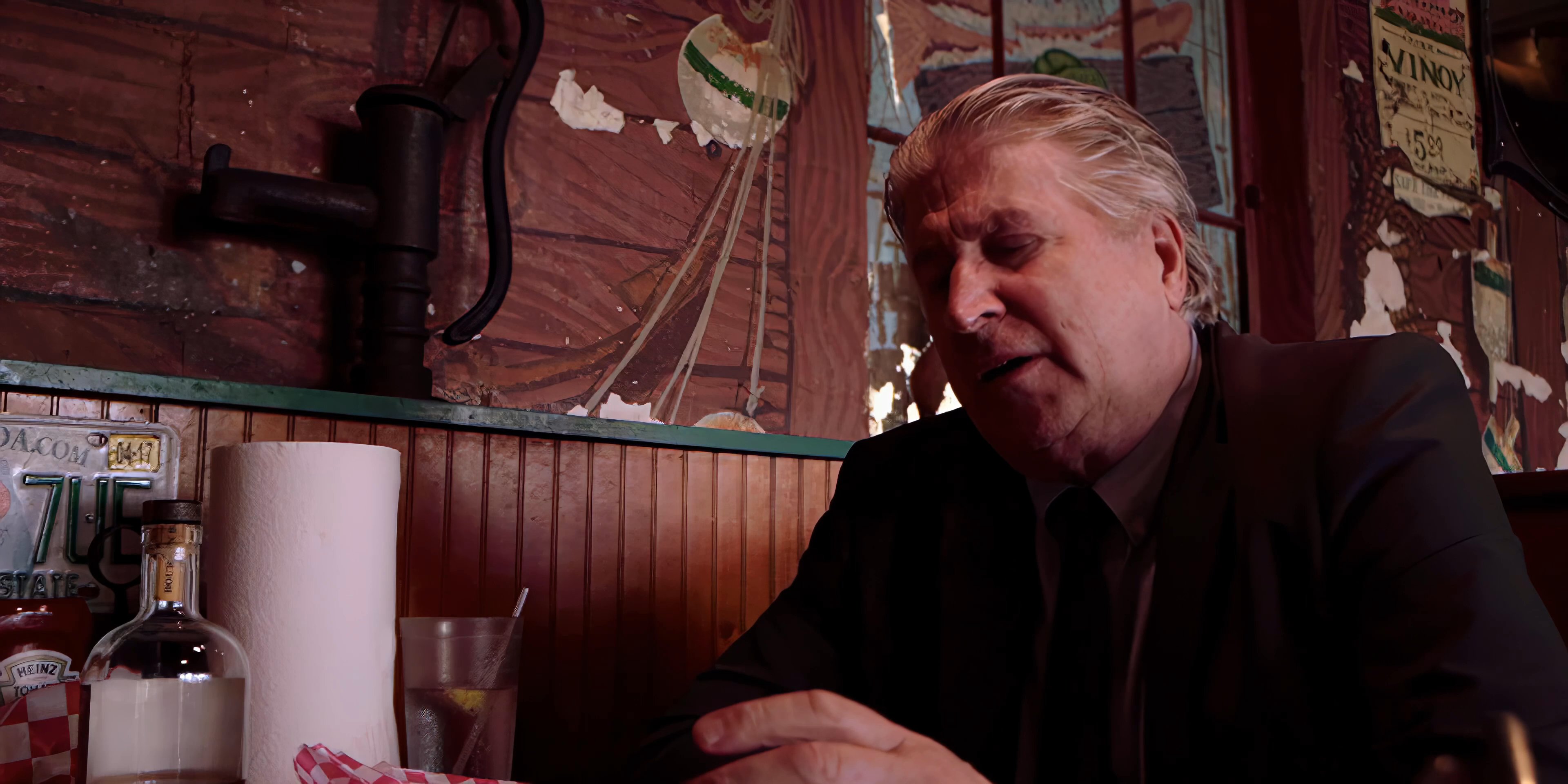 An older man in a suit engages in an animated discussion with another individual inside a diner setting. The primary speaker gestures expressively while sharing a narrative with his seated companion.