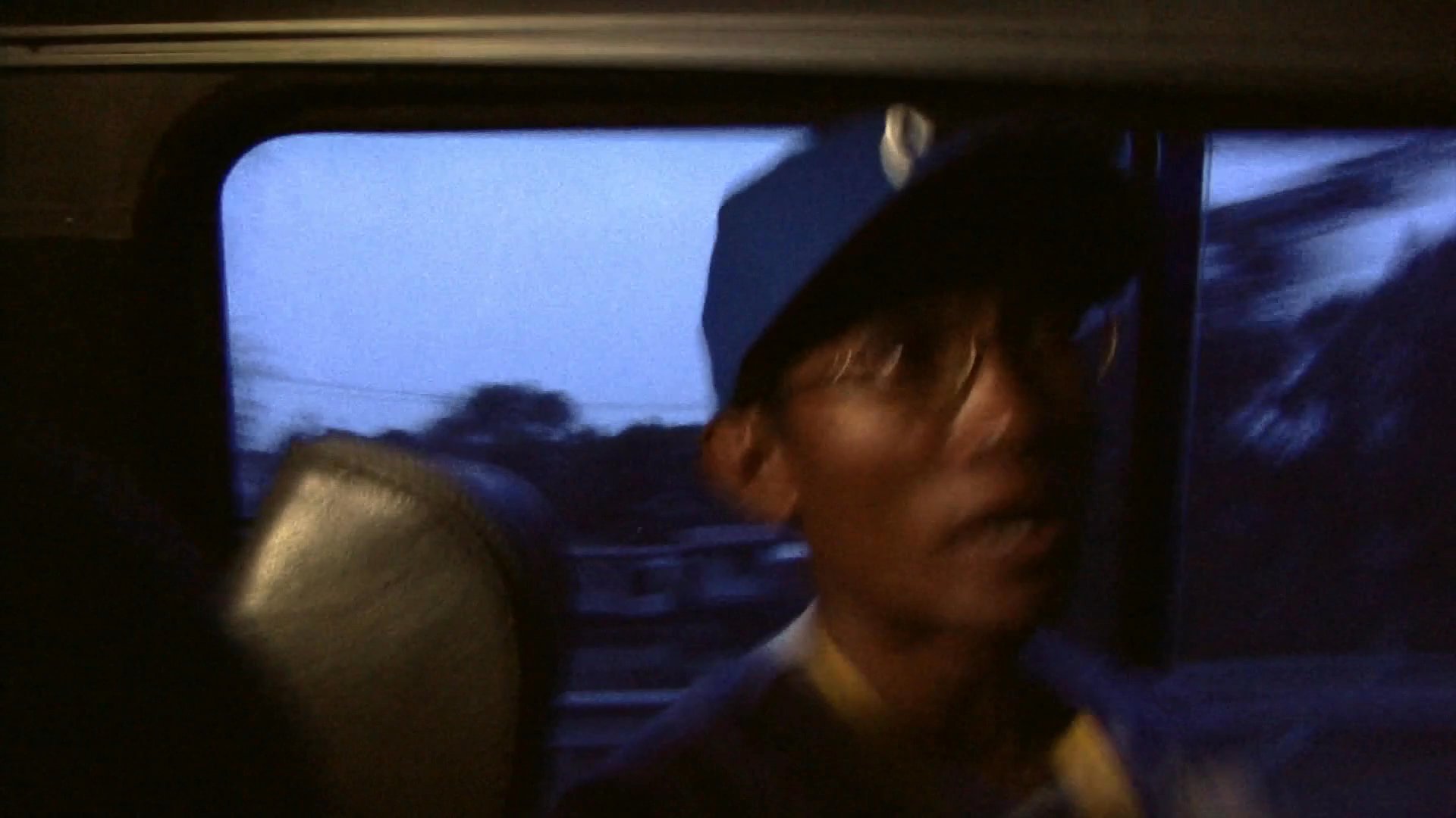 A man wearing a baseball cap sits in the passenger seat of a moving van during twilight. He speaks to an off-camera interviewer while looking out the window as the landscape blurs in the background.