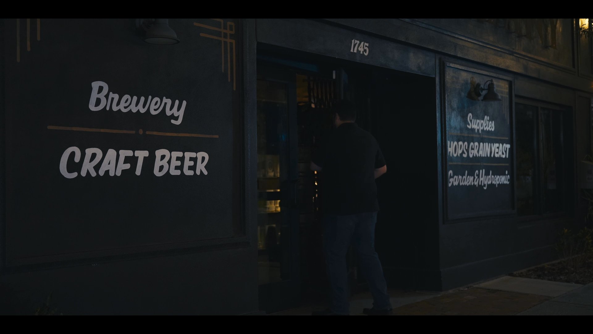 A man exits a craft brewery at night, closing the door and walking away from the establishment. The scene contrasts the cozy, well-lit interior bar area with the dark, quiet street outside.