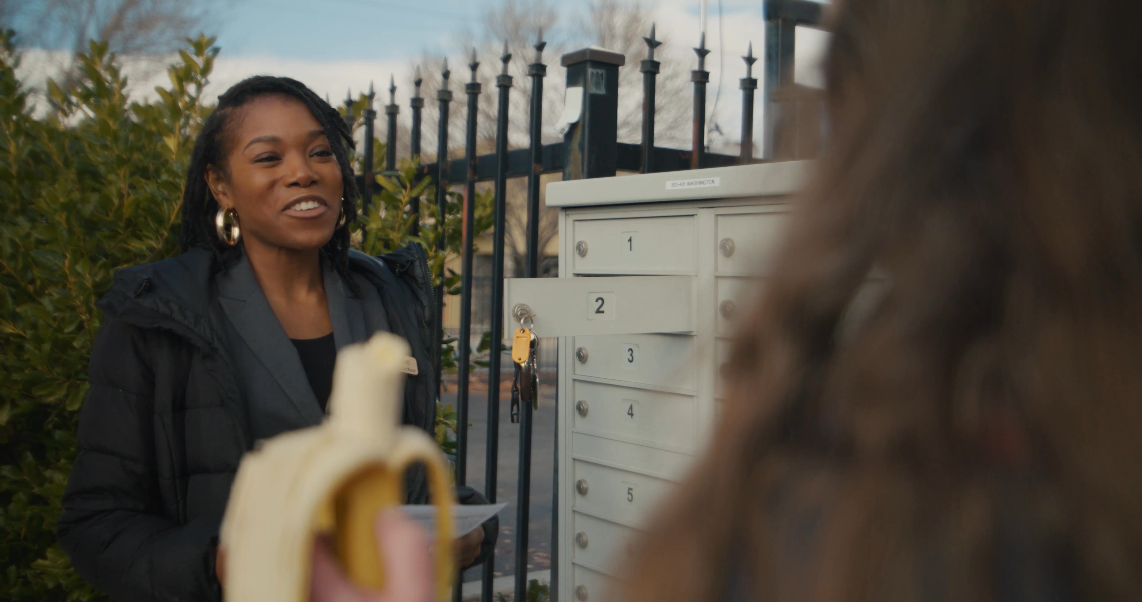 Two women engage in a casual conversation near a row of mailboxes, ultimately exchanging a bottle of prescription medication.