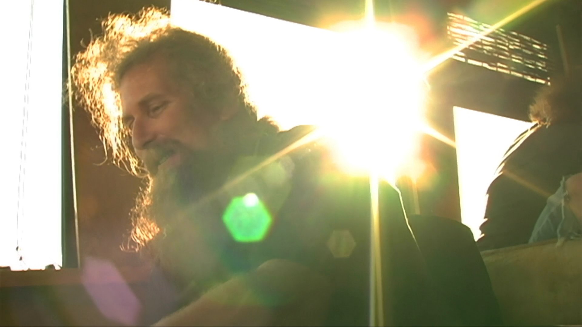 A man with a long beard recounts experiences during an interview while riding in a touring bus with companions. The scene transitions to atmospheric footage of the travelers seated inside the vehicle and observing the scenery through the windows.