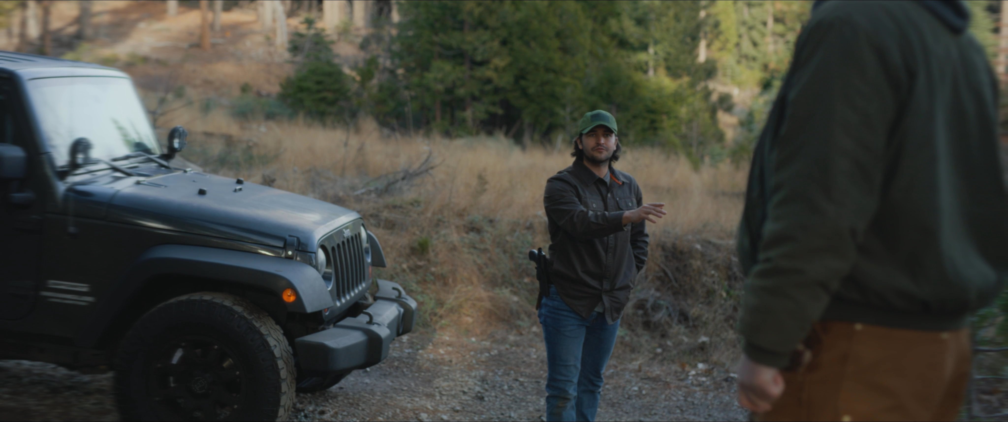 A sheriff in rugged work attire interacts with another individual near a patrol vehicle in a forested area. They appear to be discussing a situation while conducting an outdoor investigation.