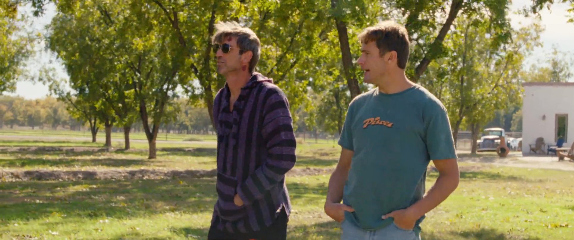Two men discuss a situation while walking through a sunny orchard, with one showing signs of a recent facial injury. The interaction maintains a serious but quiet tone as they navigate the outdoor space.