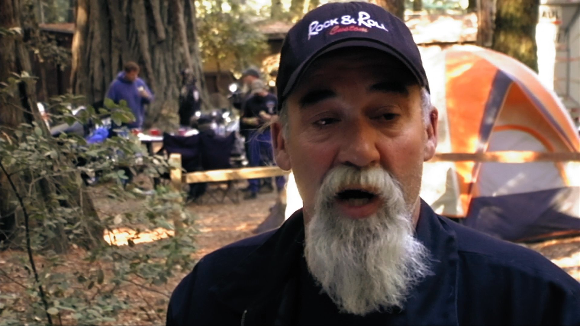 A man with a white beard and a motorcycle cap discusses his experiences while camping at a scenic outdoor location during a motorcycle trip.