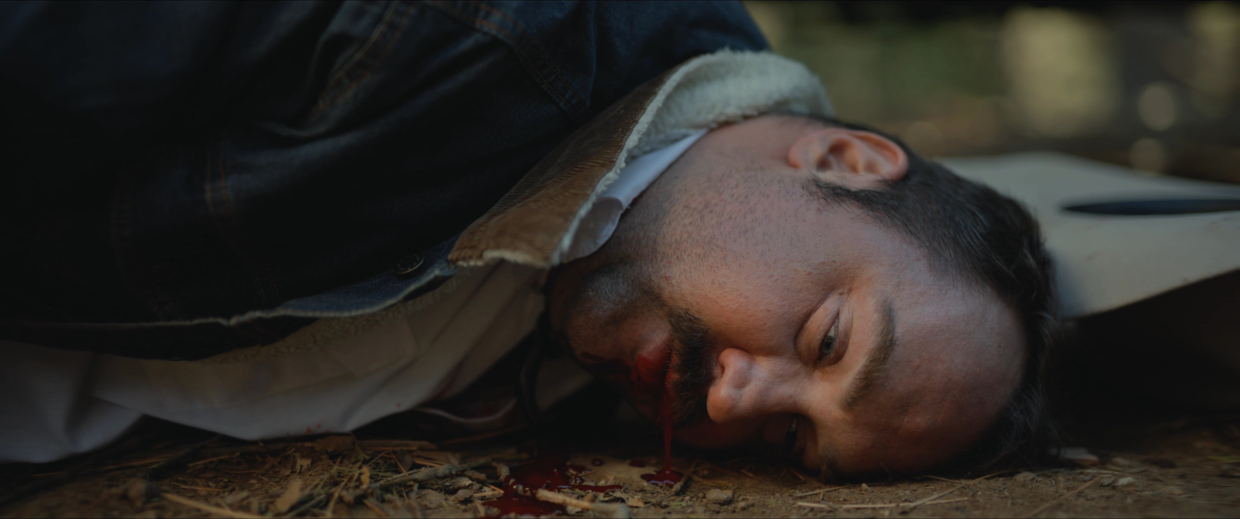 A law enforcement officer discovers a deceased individual lying on the forest floor beside a firearm and a tactical bag. The officer assesses the scene while standing near a marked sheriff patrol vehicle.