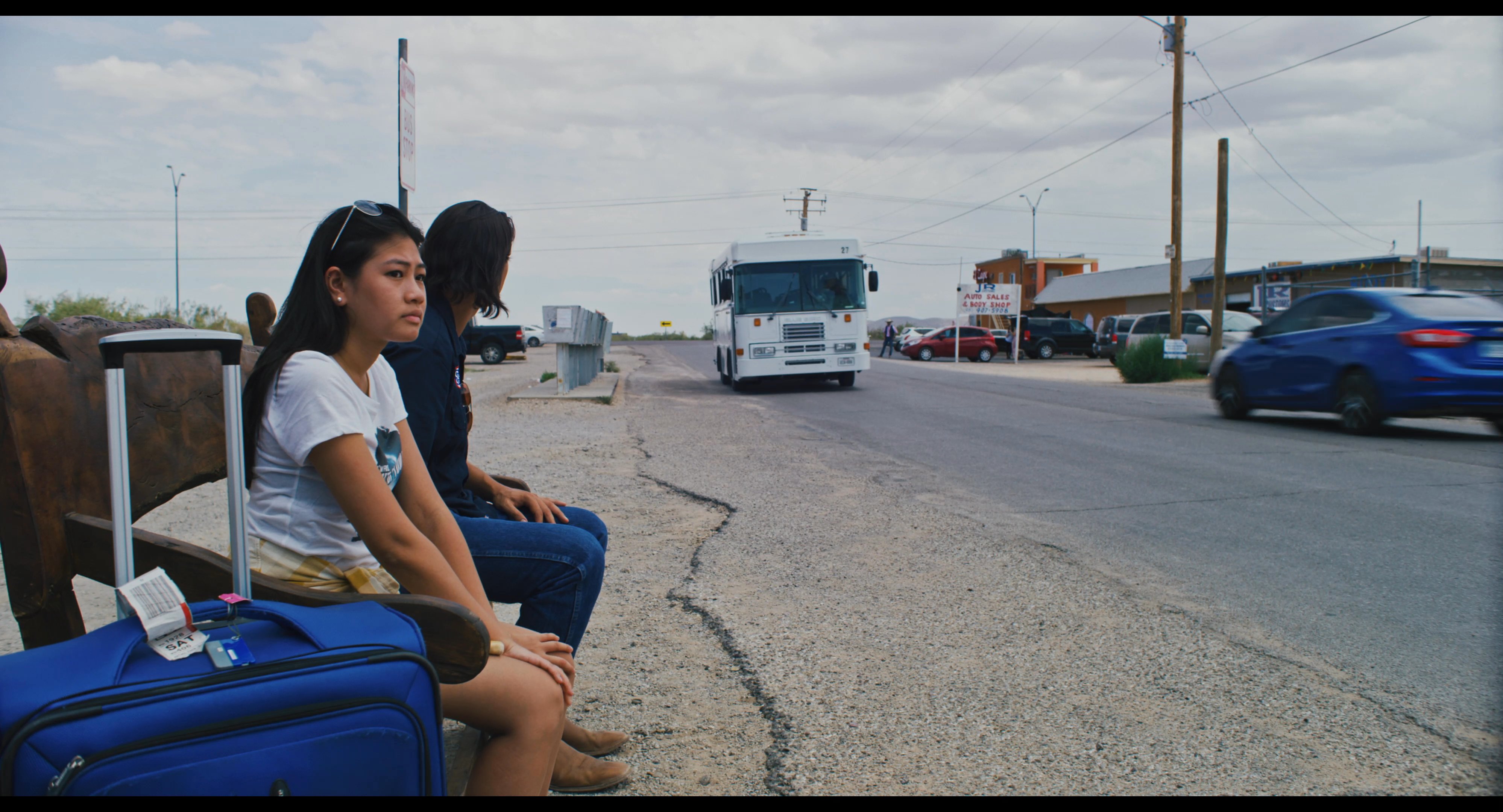 Two individuals wait on a roadside bench with luggage before a white shuttle bus arrives at their location.