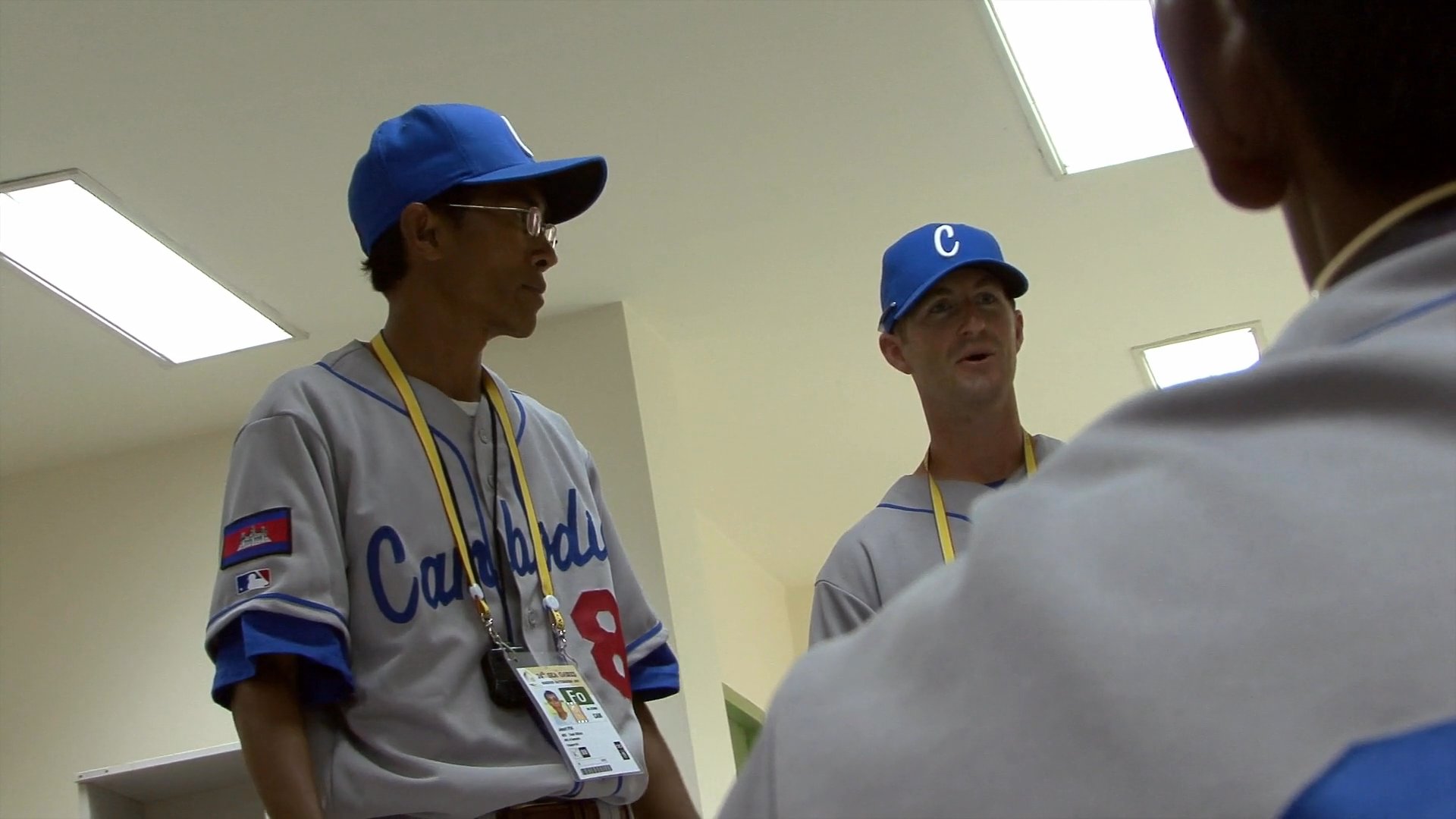 Coaches and athletes from the Cambodian national baseball team gather in an indoor space for a team briefing. The group discusses strategy and coordinates logistics while wearing team uniforms.