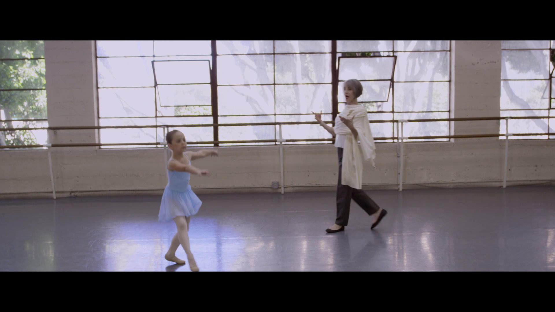 An instructor provides hands-on guidance to a young dancer, correcting her posture and movement within a professional ballet studio setting.