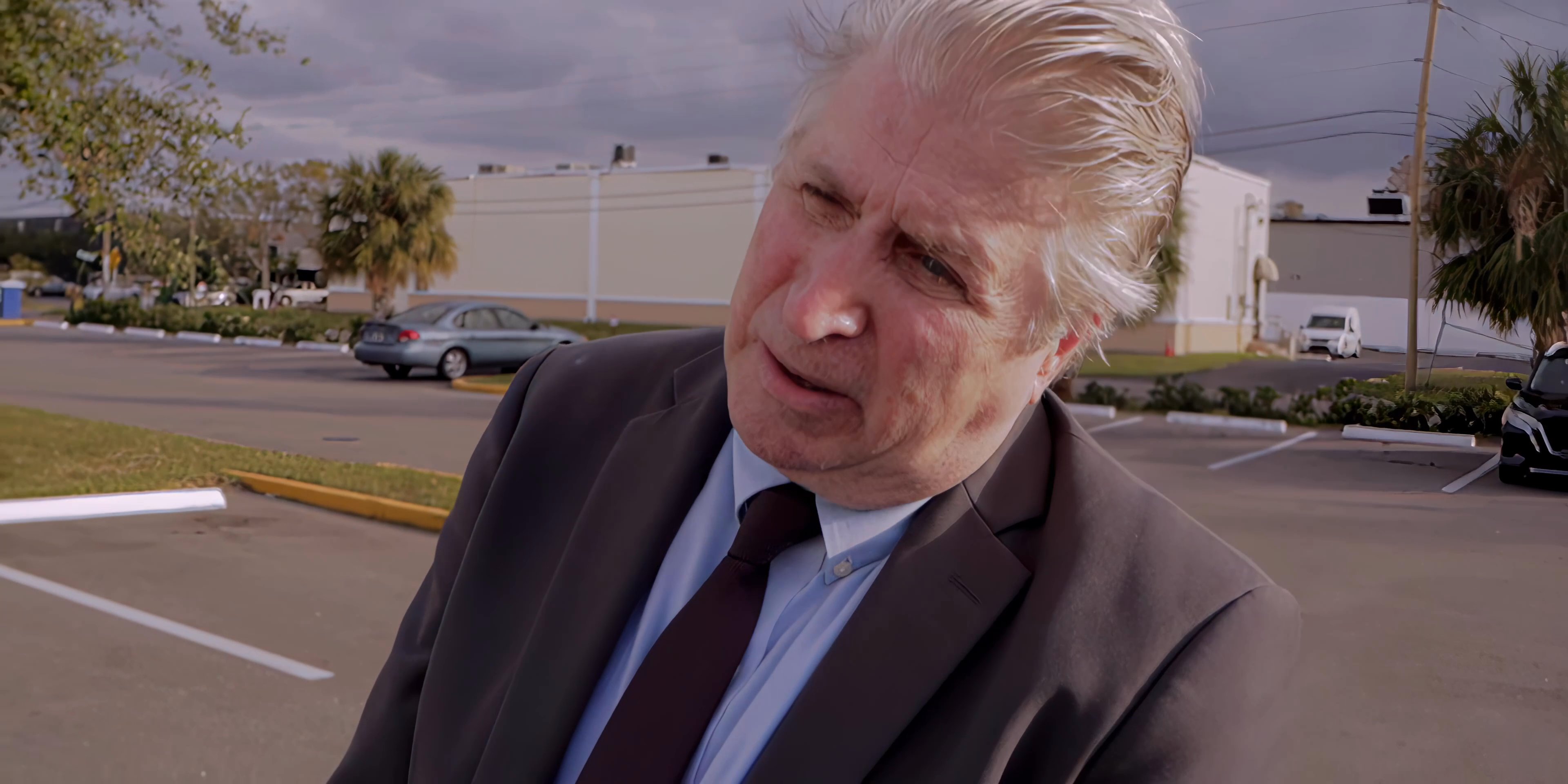 A man in a business suit and a woman in a gold top engage in a serious, focused conversation within a parking lot setting.