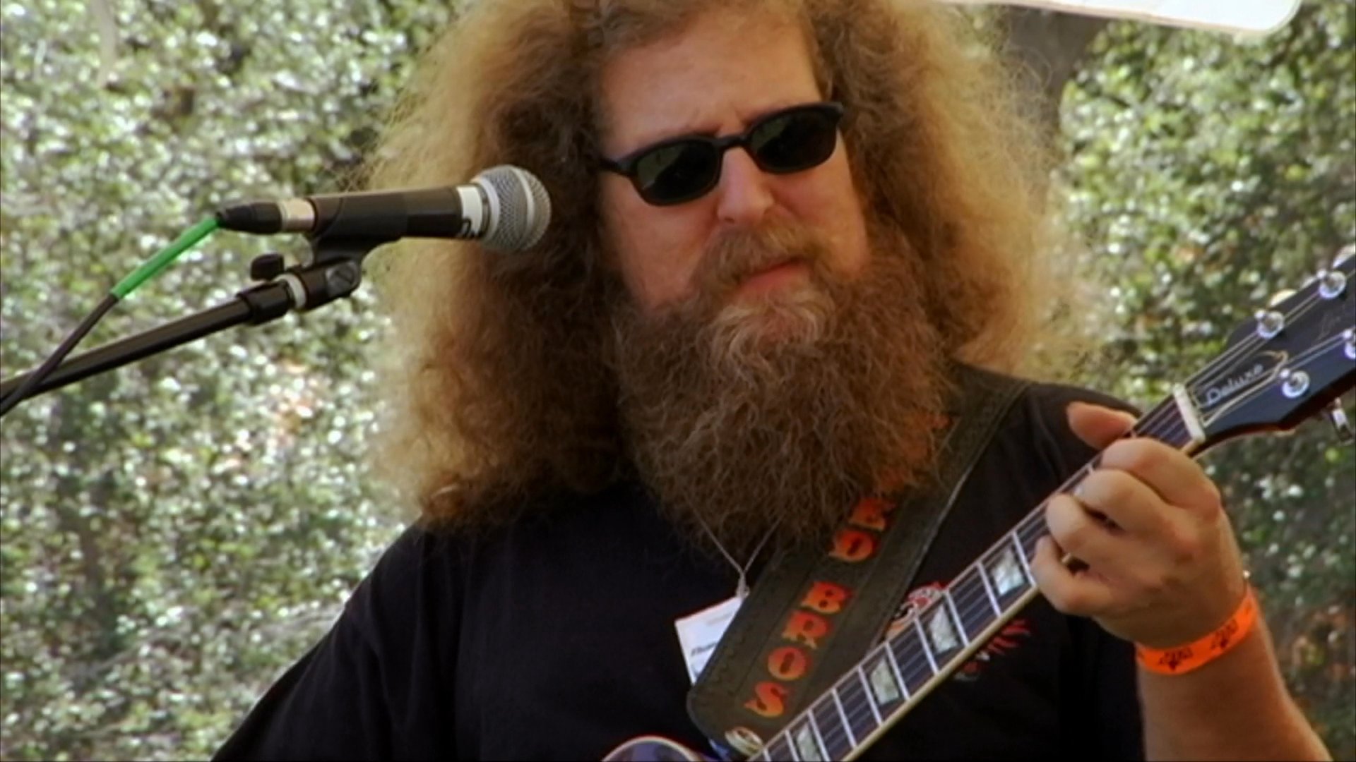 A long-haired, bearded guitarist performs live on an outdoor stage at a motorcycle event, while custom motorcycles are displayed in the surrounding area.