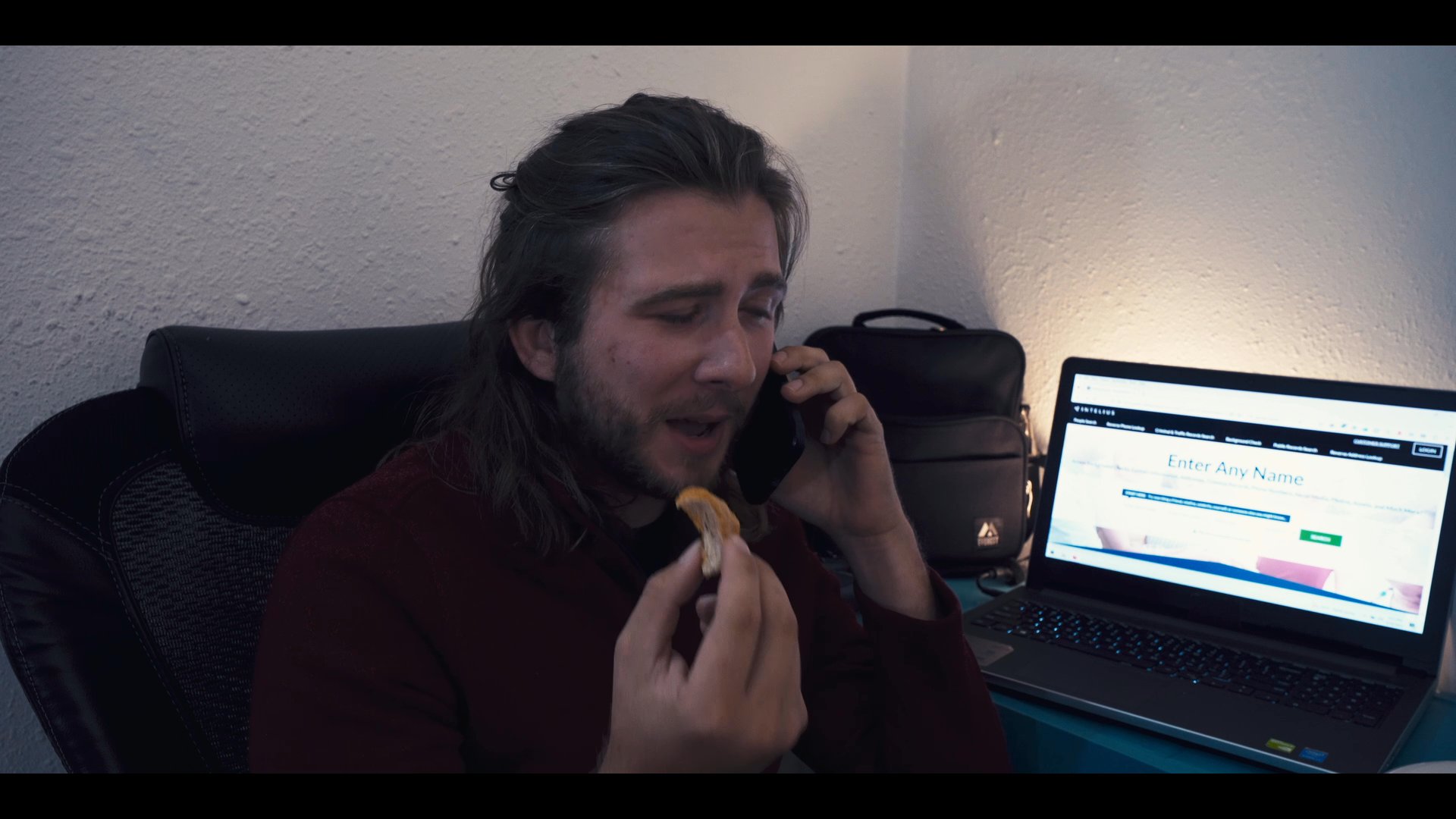 A man participates in a late-night phone call while working at a computer in a dimly lit home office. The scene alternates between his workspace and a separate room where another individual rests in bed.