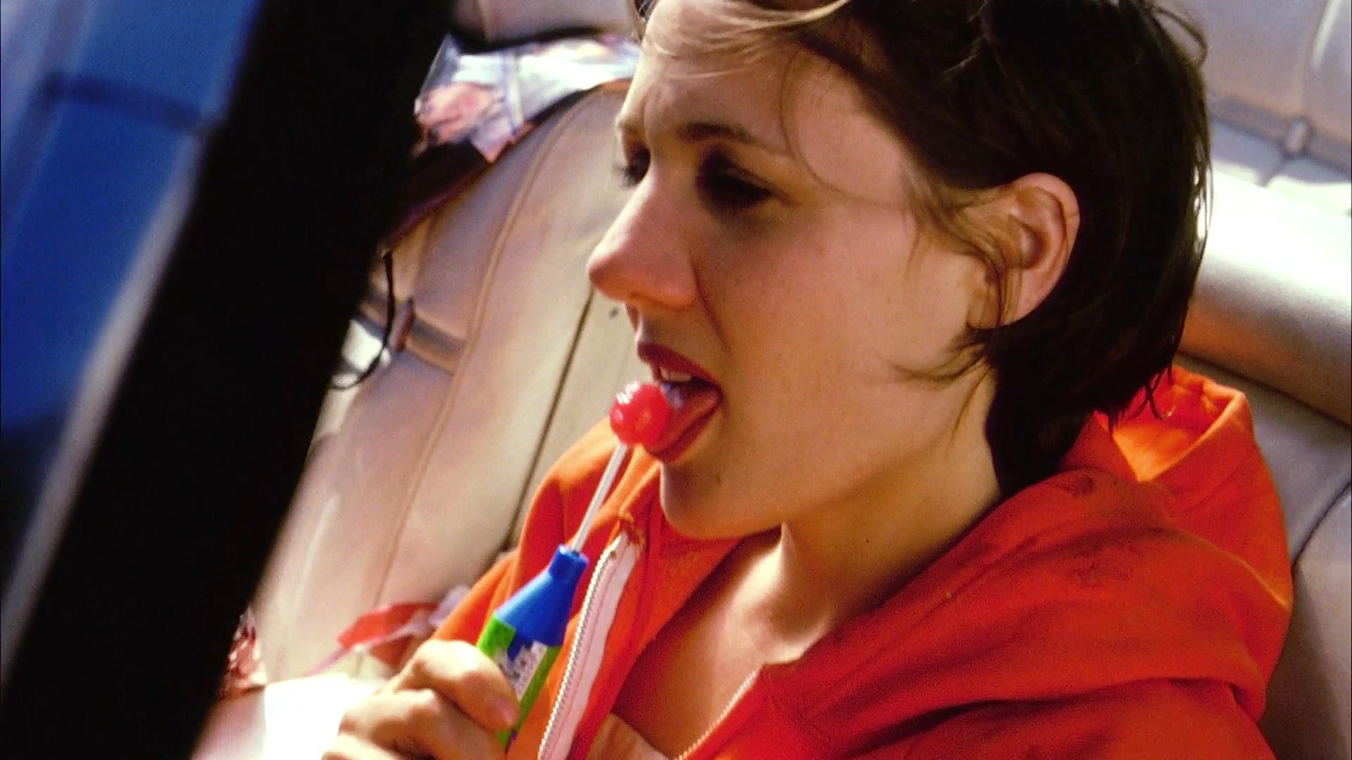 A woman drives through a rural landscape in a convertible, snacking on various food items and enjoying her solo journey.