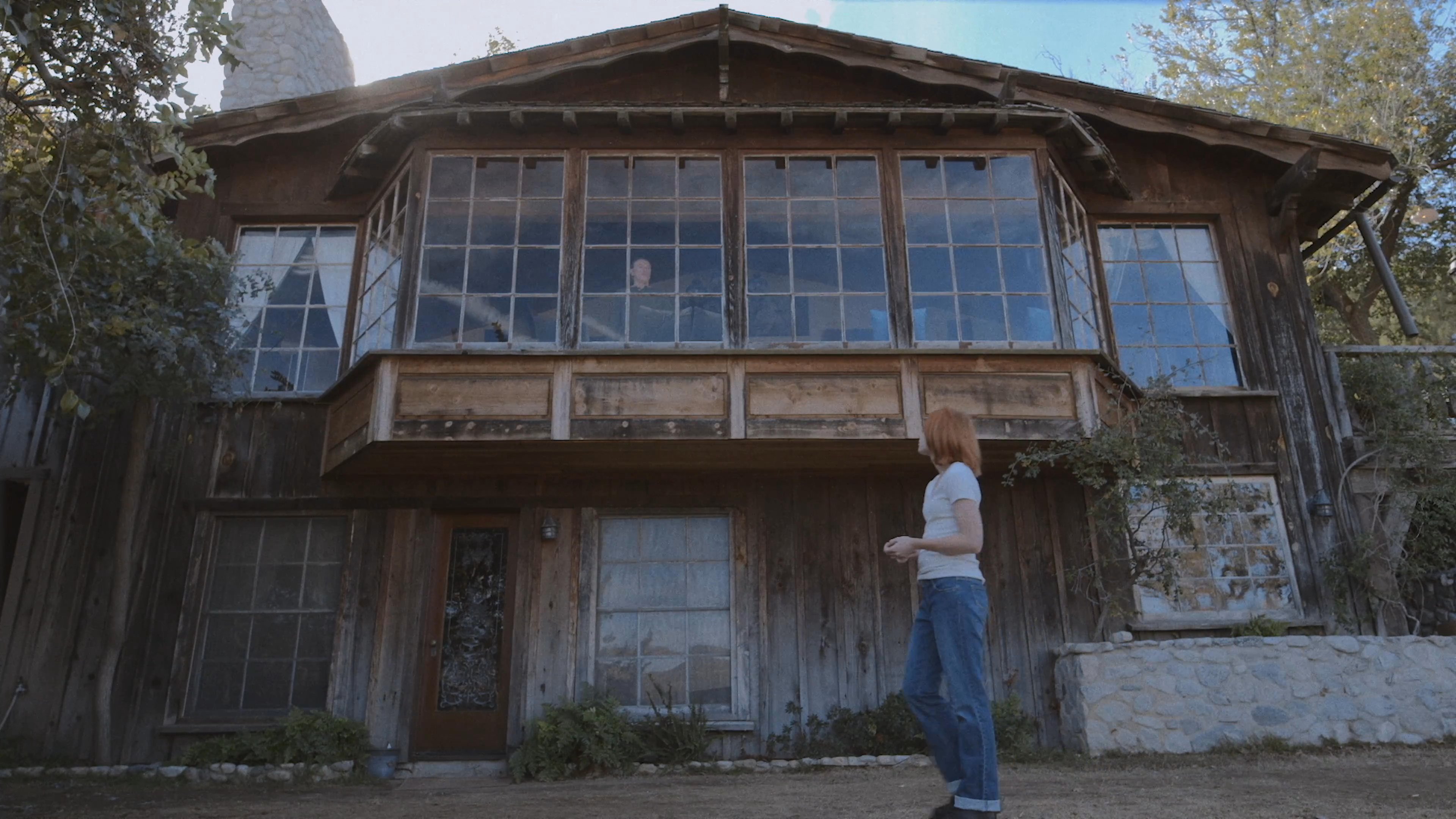A woman walks across the lawn toward a multi-story rustic cabin featuring large windows and exposed wood siding. The scene transitions to a detailed view of stained glass inside the home.