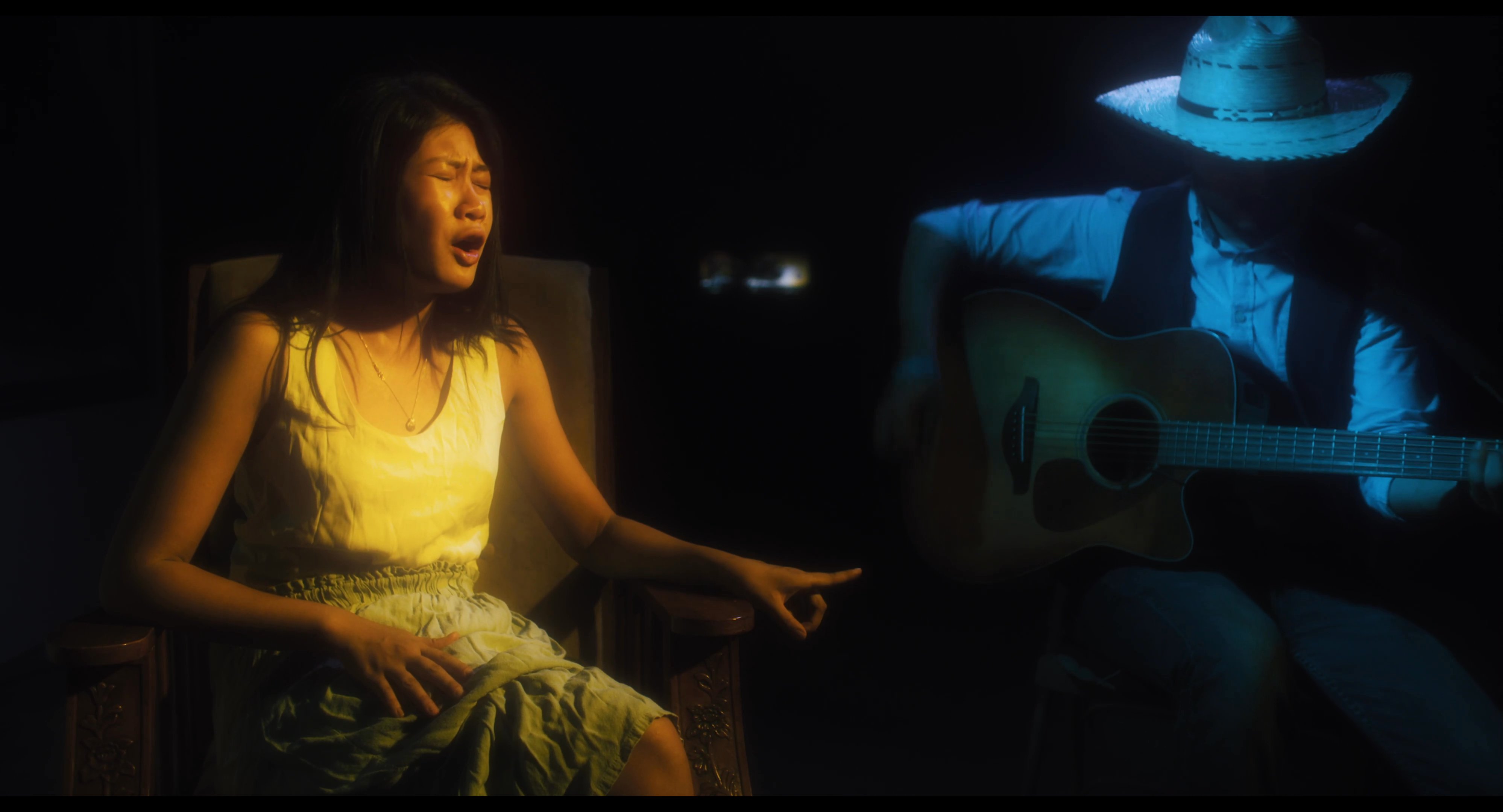 A female vocalist and a male guitarist wearing a cowboy hat perform an acoustic song in a dimly lit, atmospheric setting.