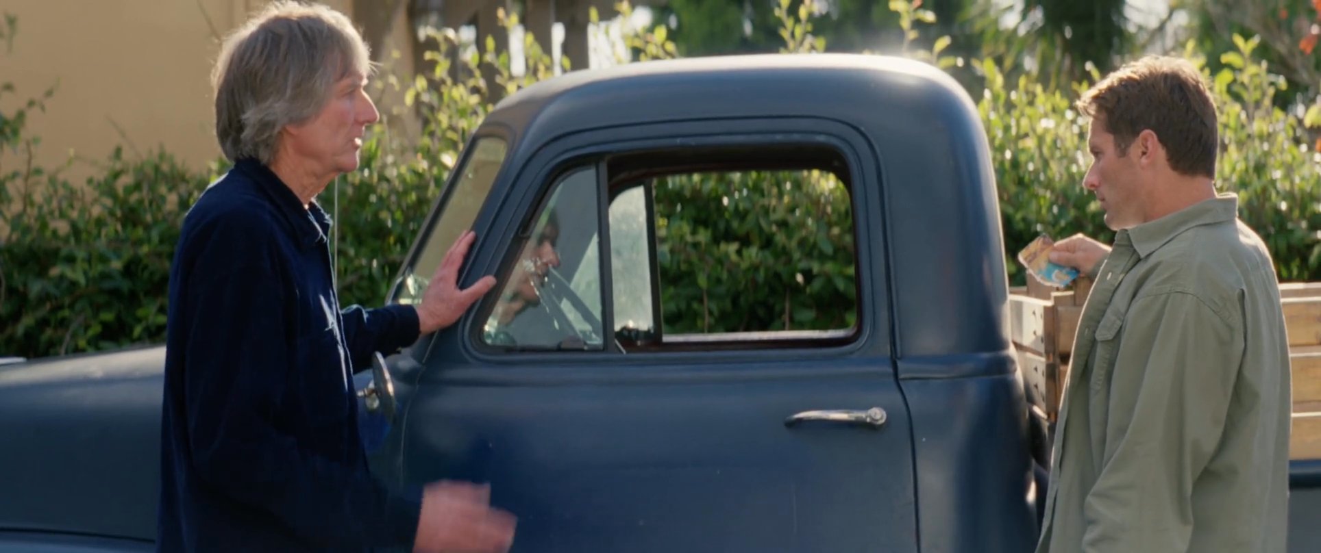 Two men engage in a professional discussion beside a vintage blue pickup truck before finalizing their agreement with a handshake. The outdoor setting provides a neutral backdrop for their interaction.