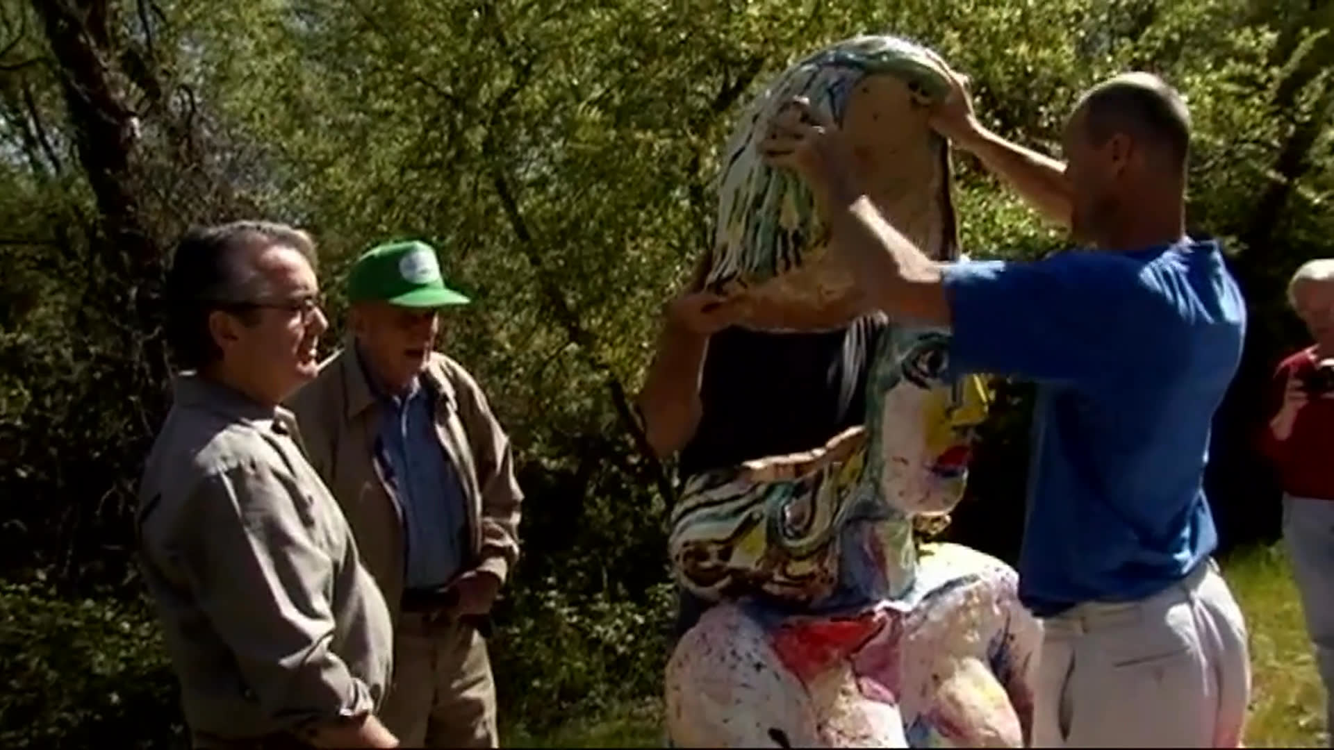 Jack Rasmussen discusses the travelability of large artworks, including a sprawling twig-nest sculpture. Visitors handle a colorful head sculpture outdoors before the sequence moves inside to sculptures displayed in a gallery.