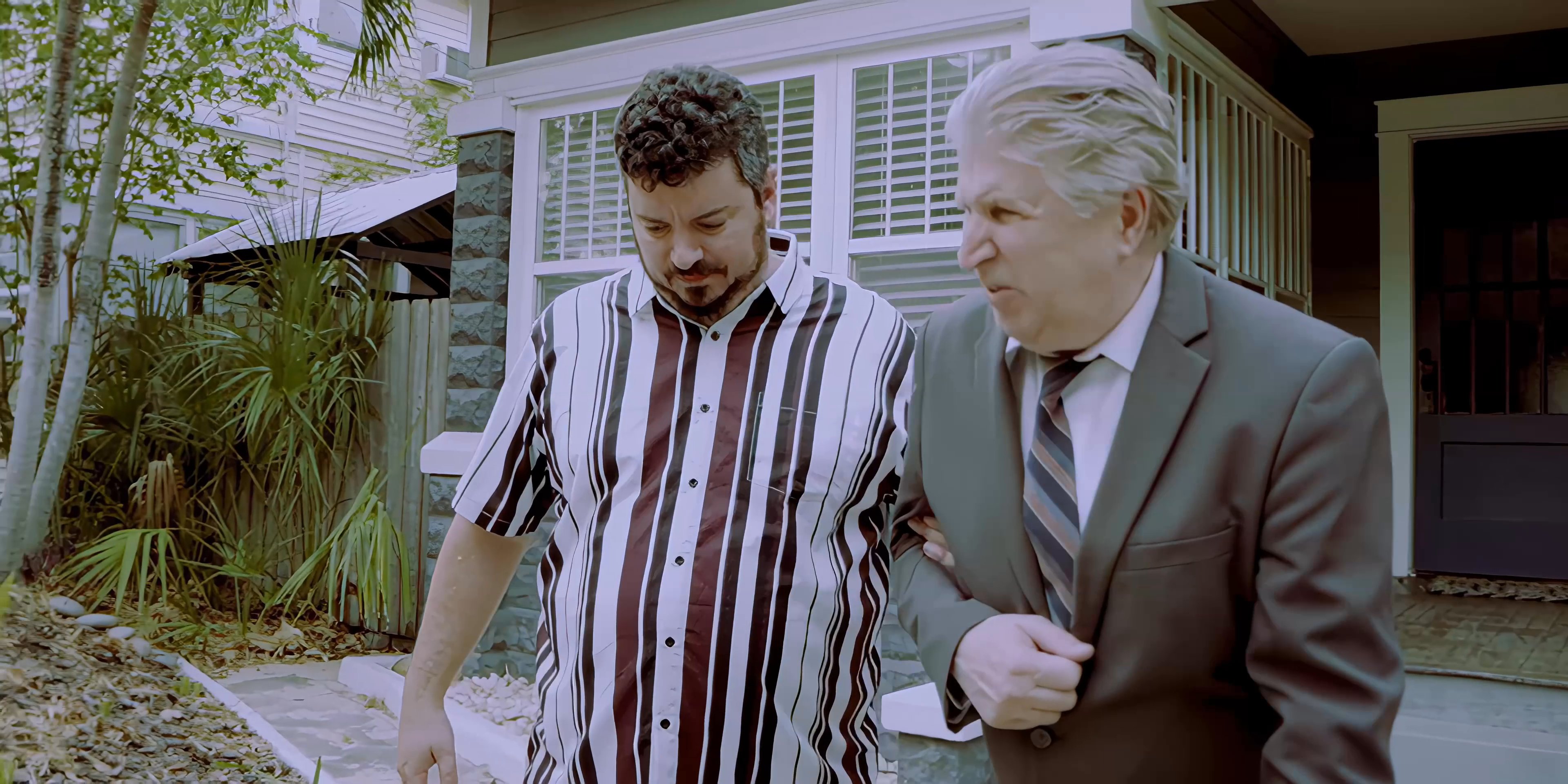 A man provides physical support and guidance to an older gentleman as they walk together along a residential pathway. The pair maintains a steady, deliberate pace while engaging in conversation.