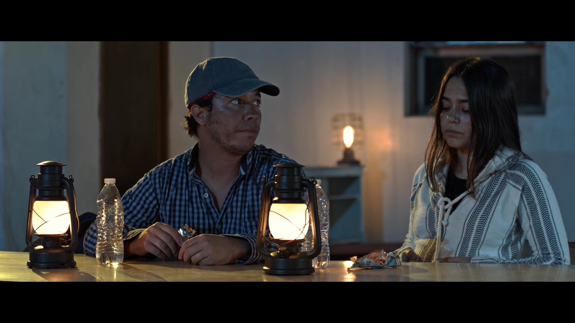 A young couple sits at an outdoor table with a lantern when a priest approaches to ask if he may join them for a conversation. The exchange occurs in a quiet, dimly lit rural setting at night.