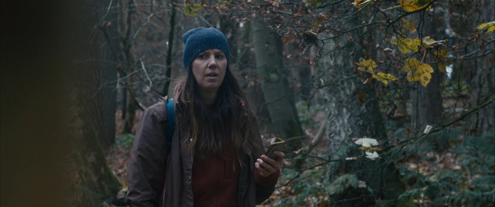A hiker navigates through a dense forest while periodically checking her smartphone for guidance. She continues her trek through the foliage, relying on her device to maintain her course.