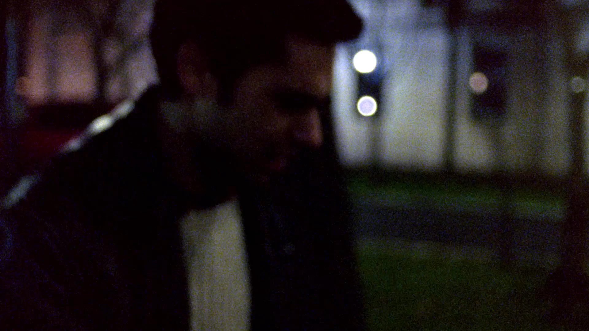 A man wearing a leather jacket and white sweater walks across a grassy area at night near a building complex. He moves steadily through the dimly lit outdoor environment.