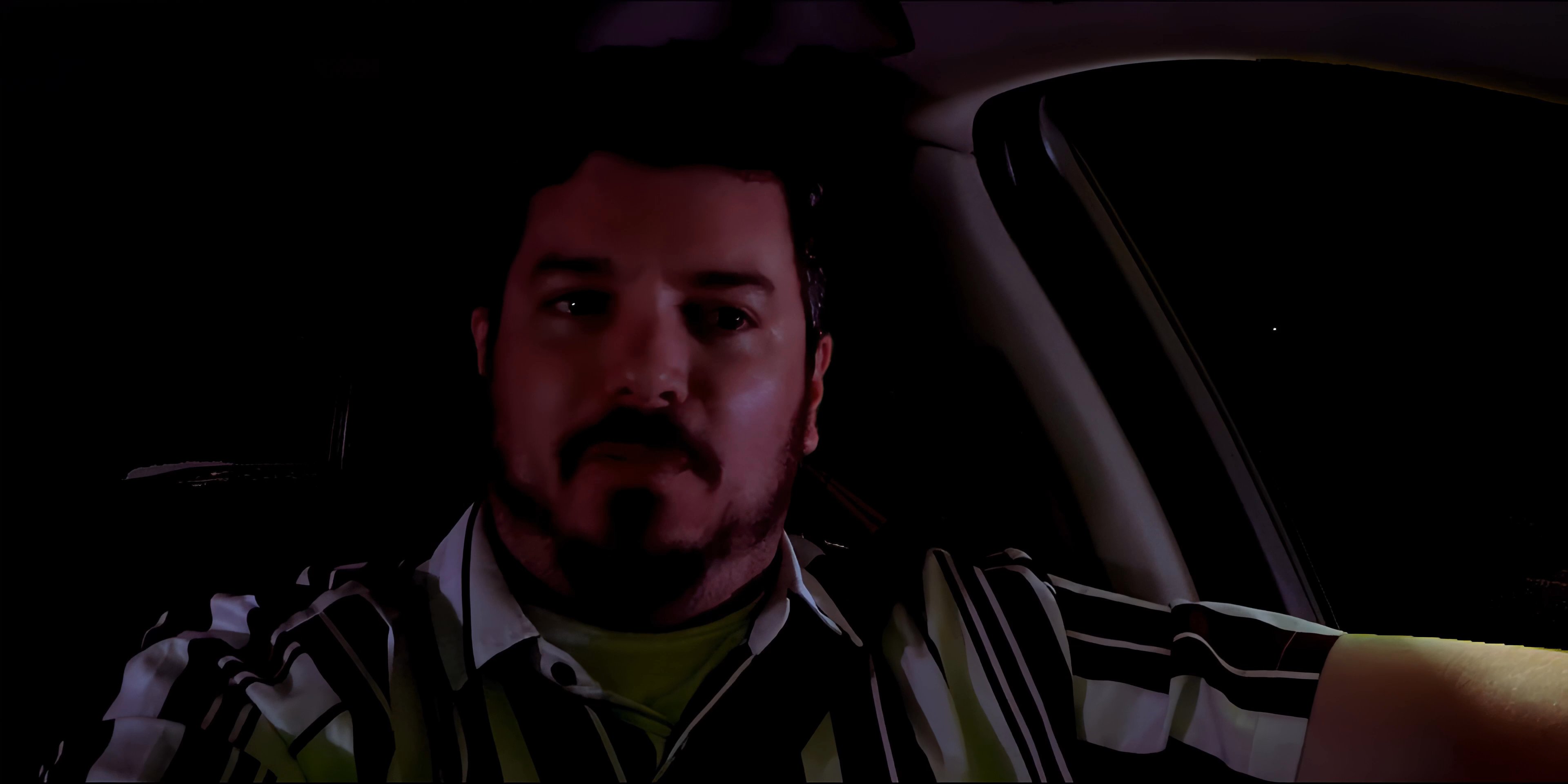 A man with a beard sits in the driver's seat of a vehicle at night, engaged in conversation while looking toward the passenger side.